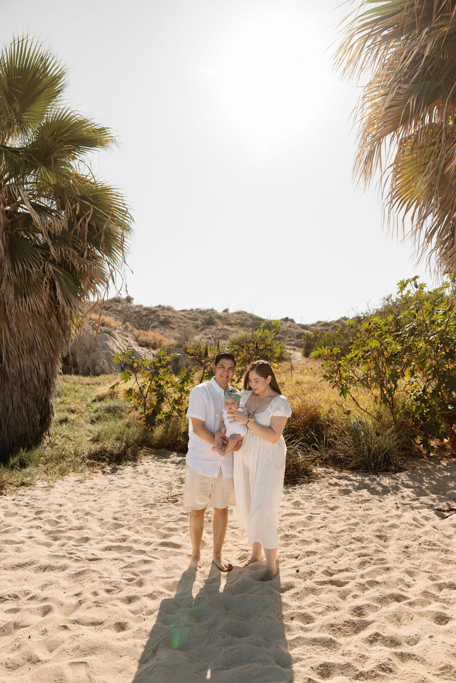 A couple in white outfits stands on sandy beach path flanked by palm trees, holding a baby in sunny coastal setting.
