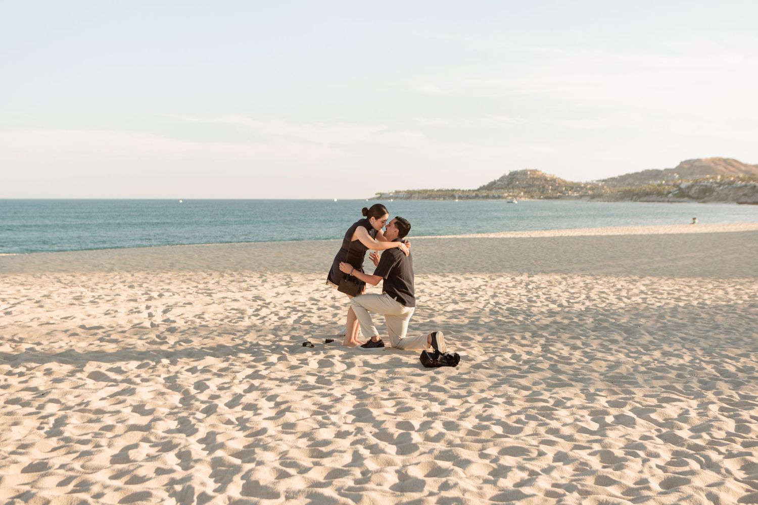 Couple sharing a romantic kiss during a beach proposal on a sunny sandy shore with ocean views.