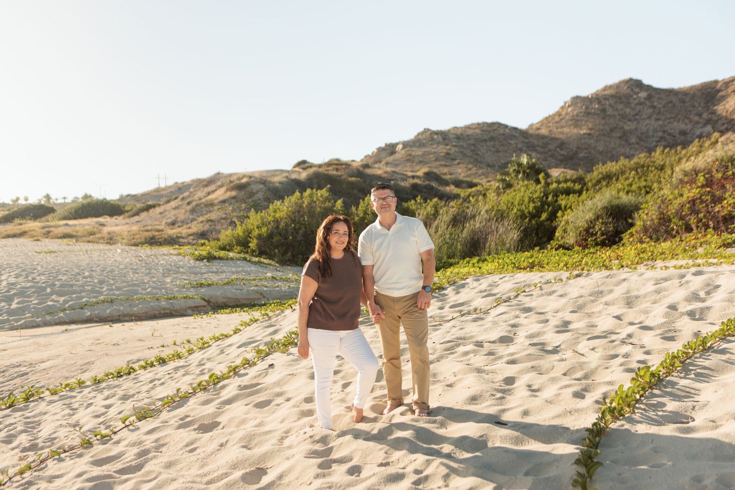 Mature couple smiles and walks hand in hand on a sandy beach path with green hills behind them.