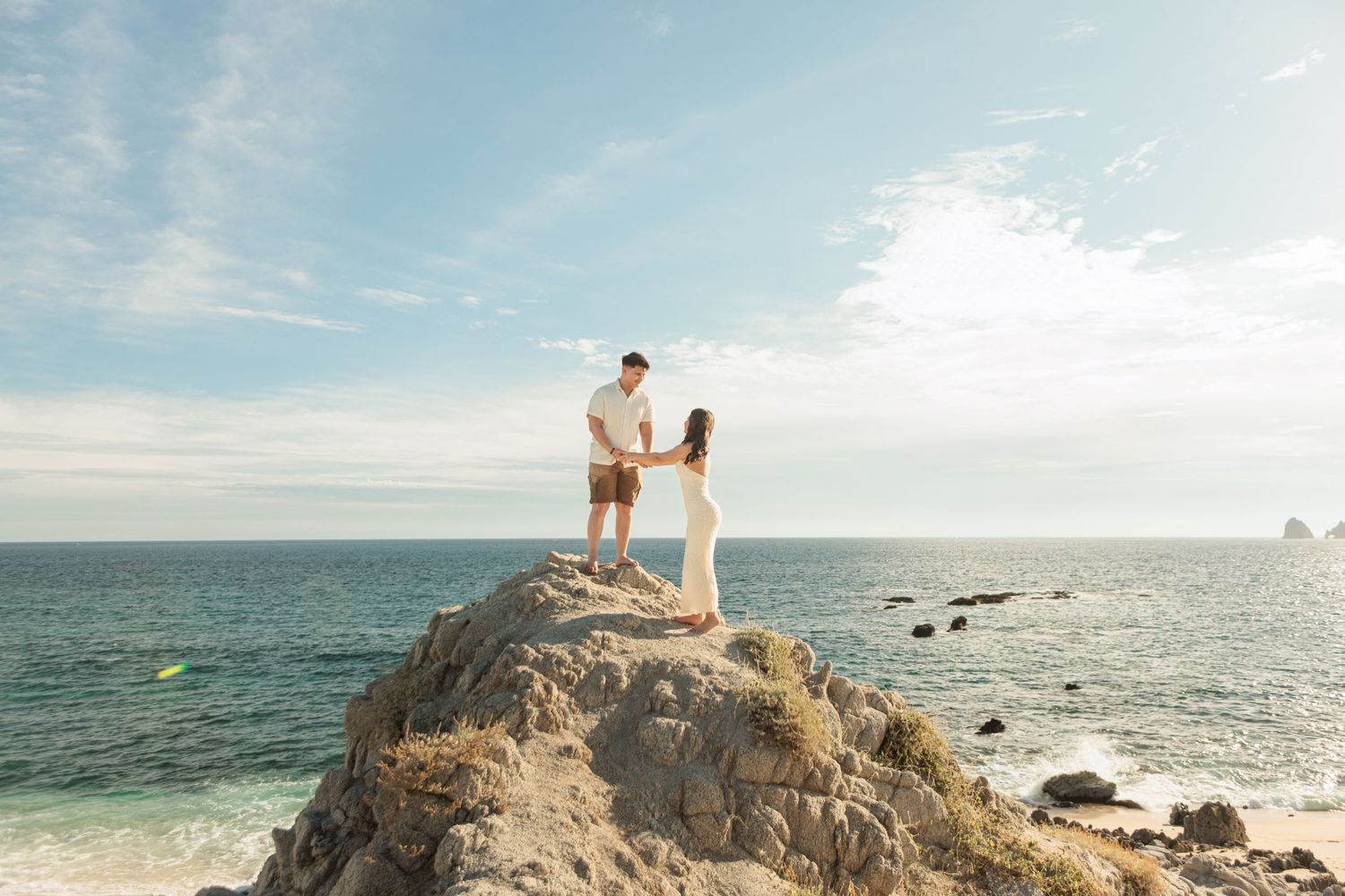 Couple holding hands on a rocky outcrop overlooking the ocean on a sunny beach day.