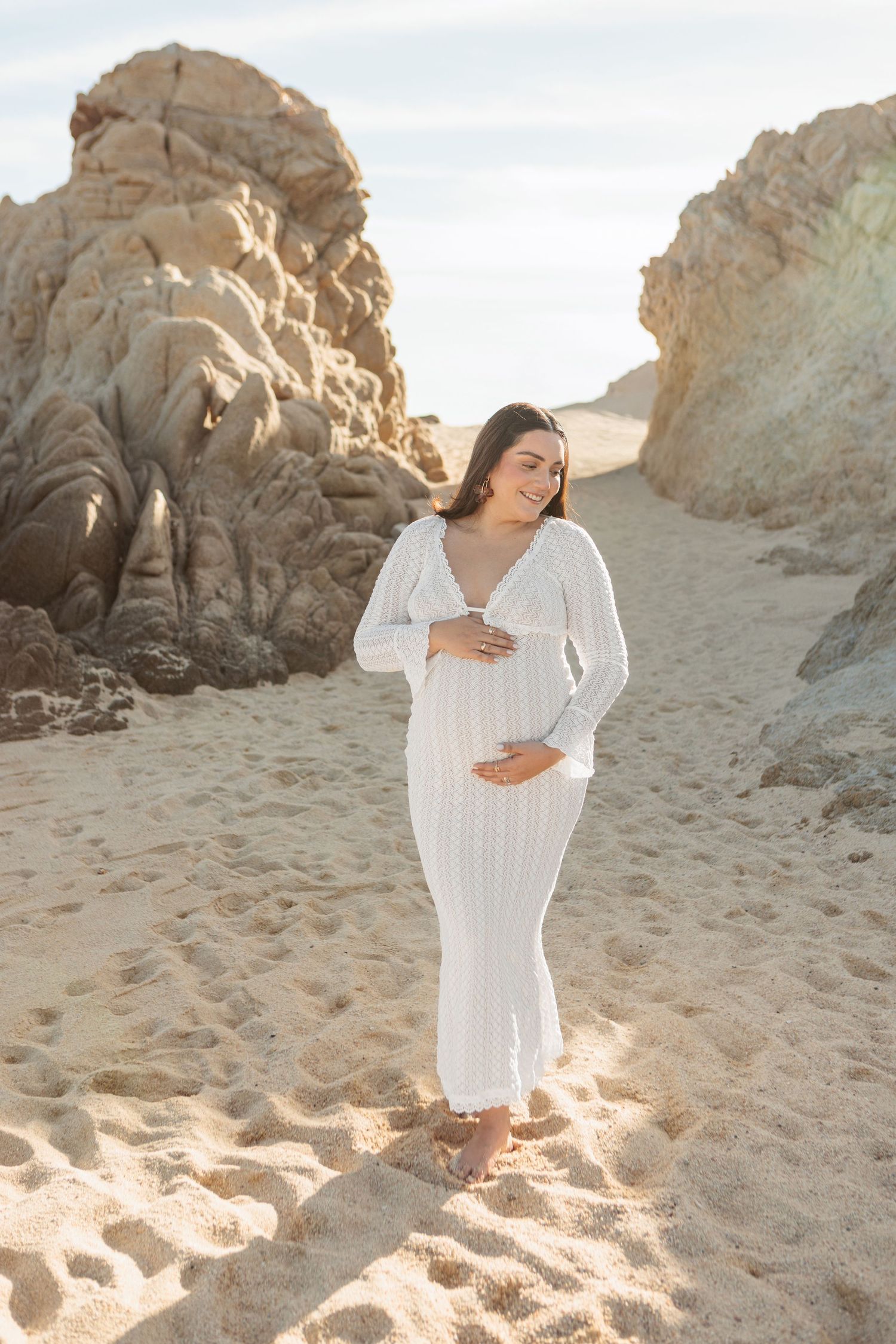 Pregnant woman in white dress cradling baby bump on sandy beach surrounded by rocky formations.