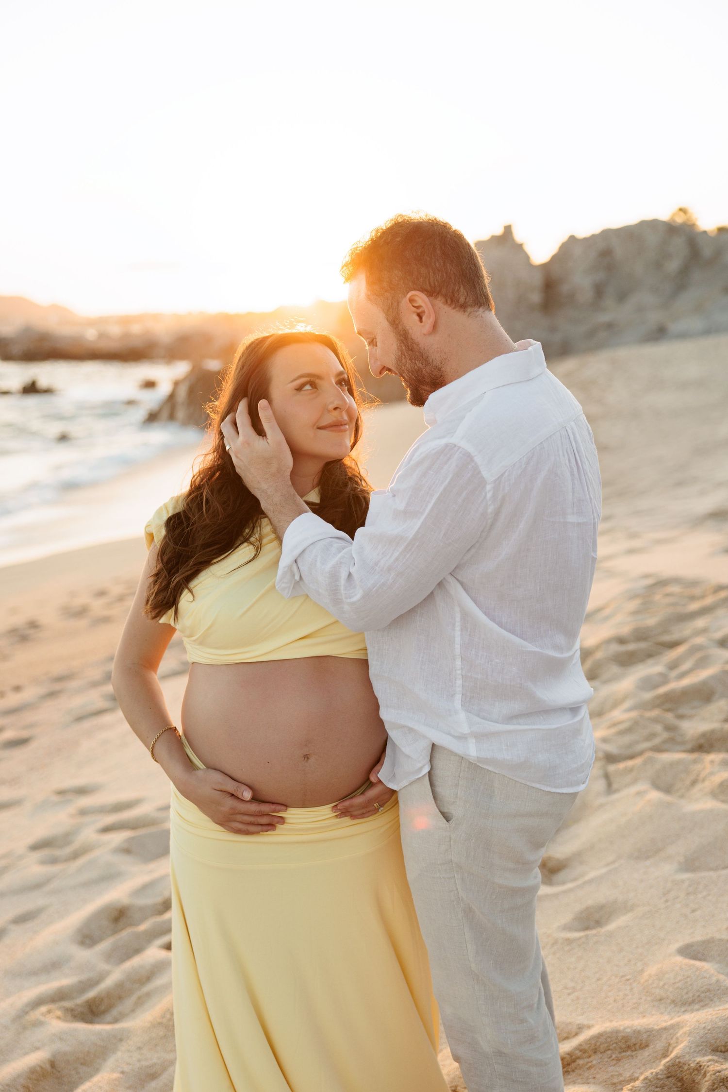 Couple sharing tender moment at sunset on beach, man gently touches pregnant womans face as she smiles.