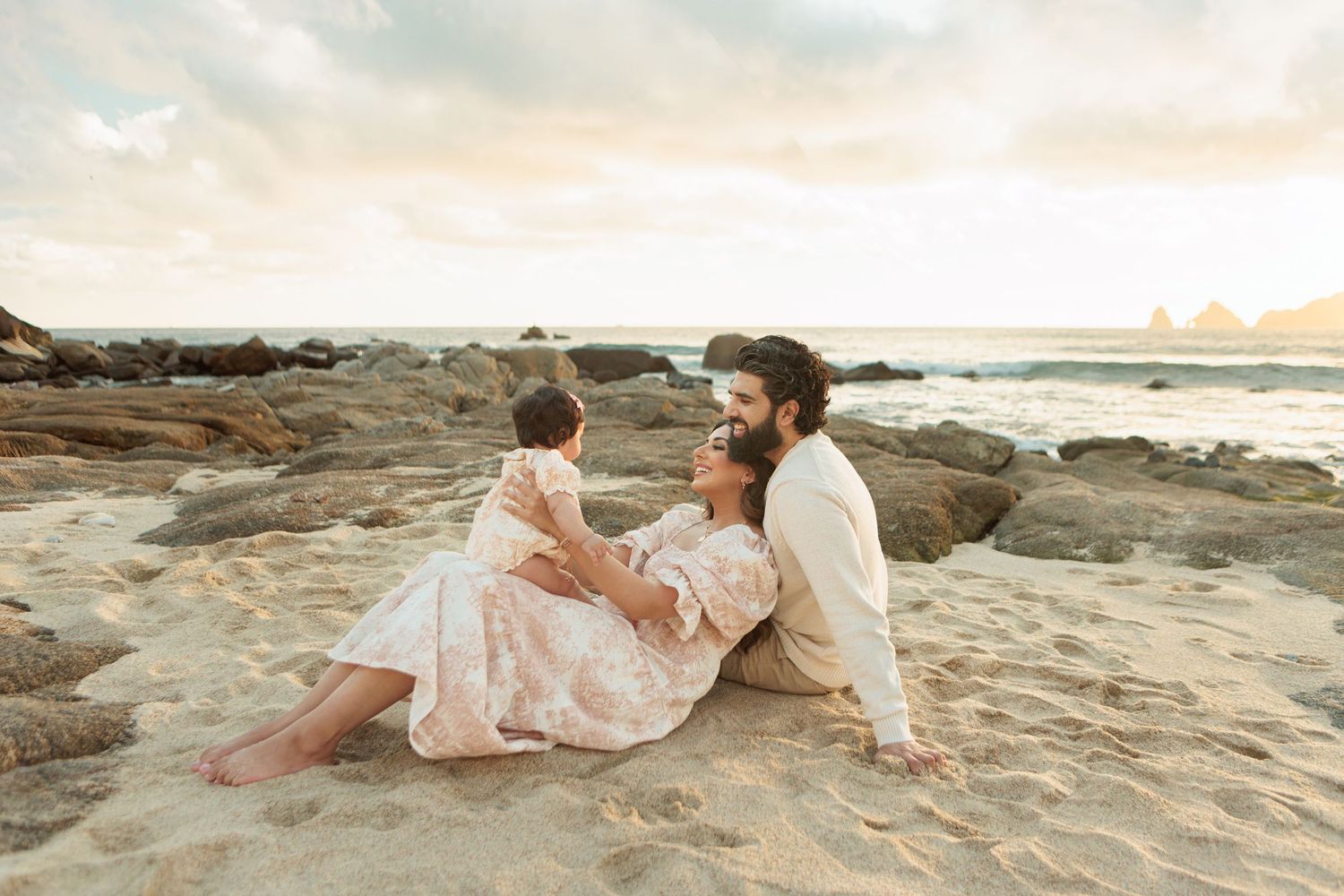 Couple sitting on sandy beach at sunset, lovingly holding their baby in a floral dress.
