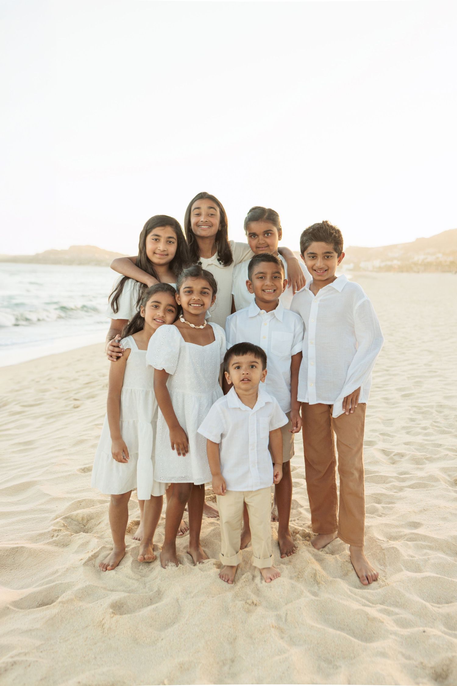 Group of eight children in white outfits posing together on sandy beach near ocean waves.