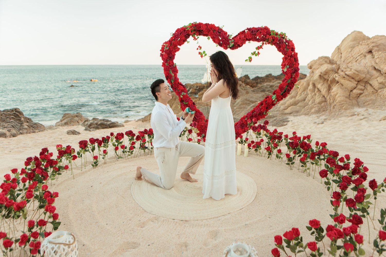 Man proposing on one knee inside red rose heart arch arrangement on beach with ocean rocks in background.