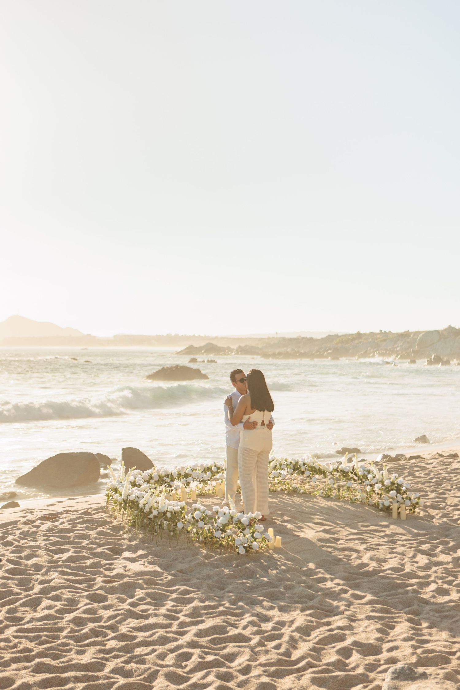 Couple embracing on sandy beach at golden hour beside white floral heart arrangement with ocean waves behind.