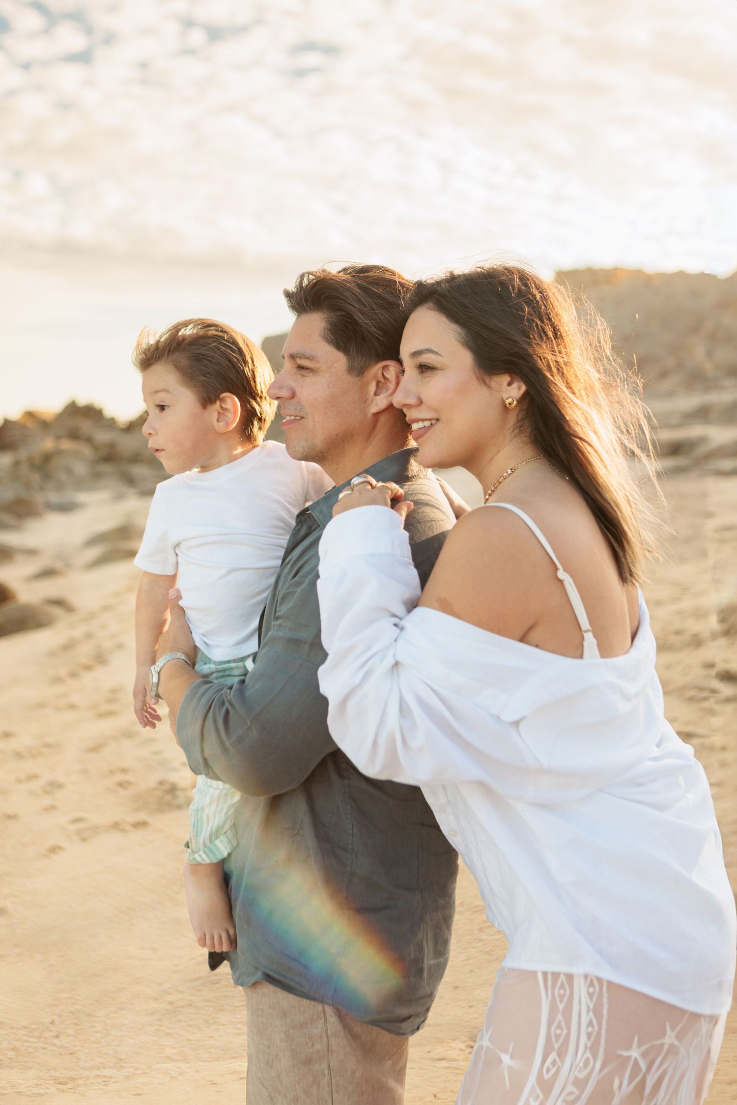 Family of three enjoying a golden sunset beach walk, father carrying young son with mother beside them.