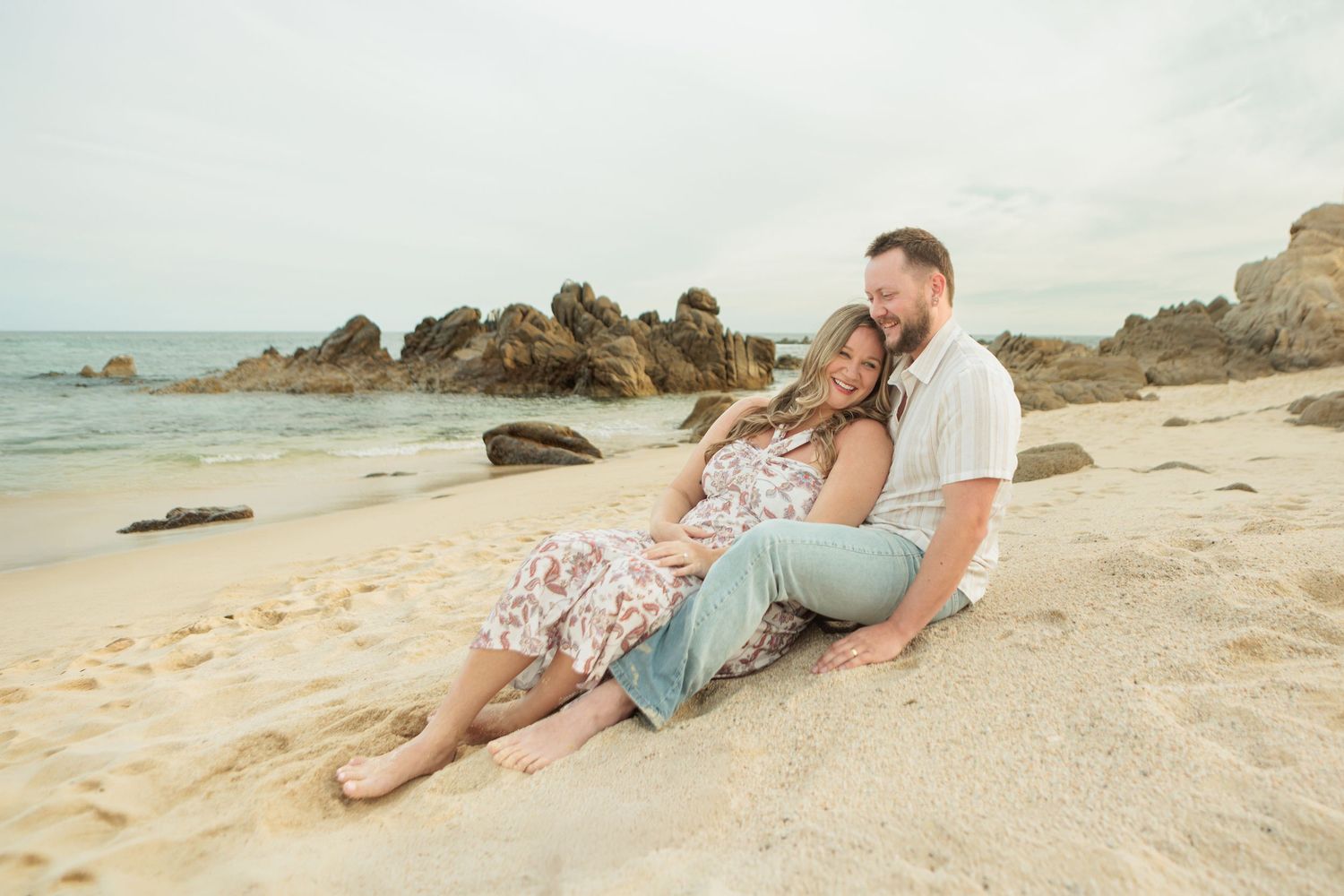 Happy couple sits together on sandy beach, woman in floral dress leans on mans shoulder near rocks.