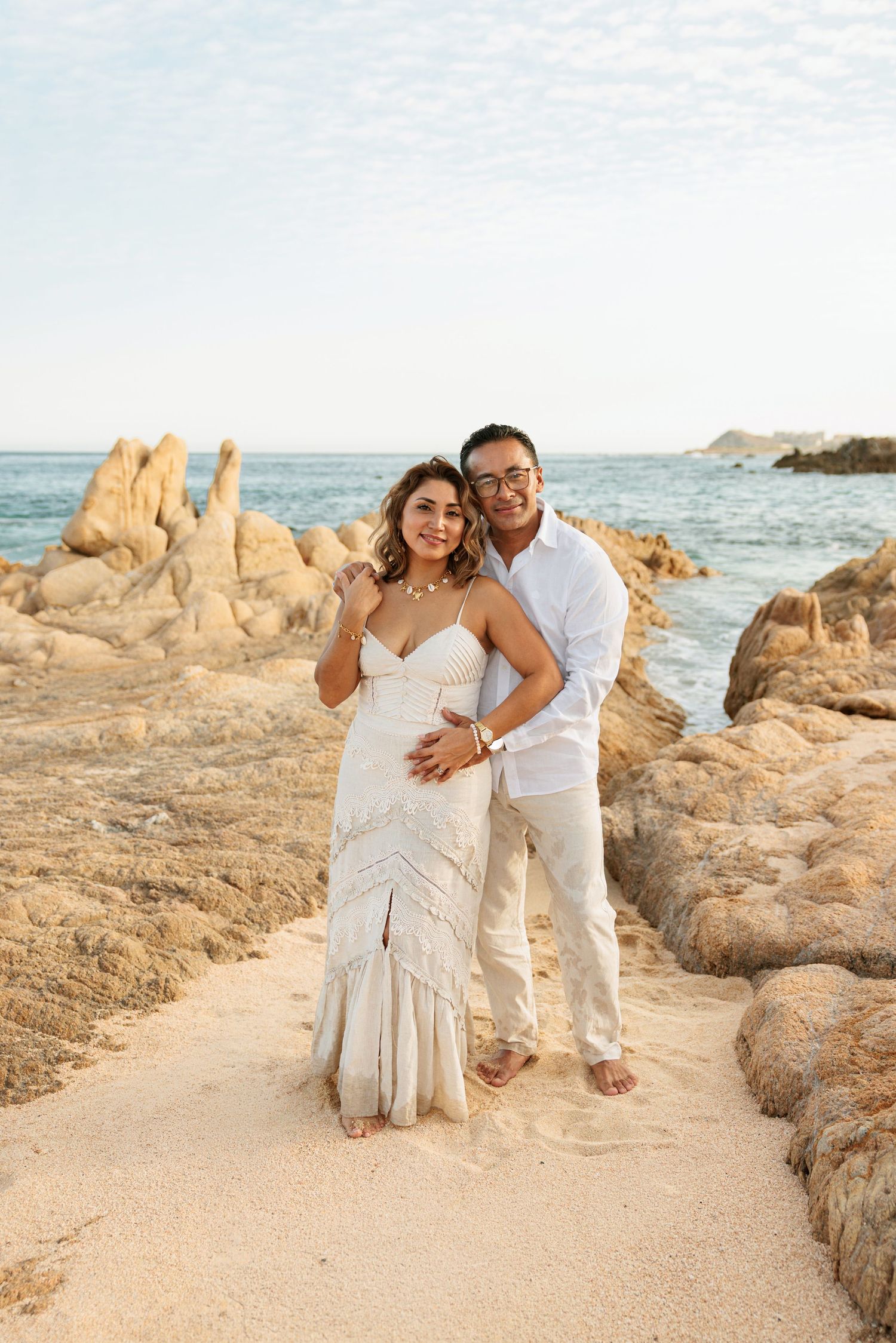 Couple posing on sandy beach path surrounded by rocky formations with ocean backdrop, wearing white outfits.