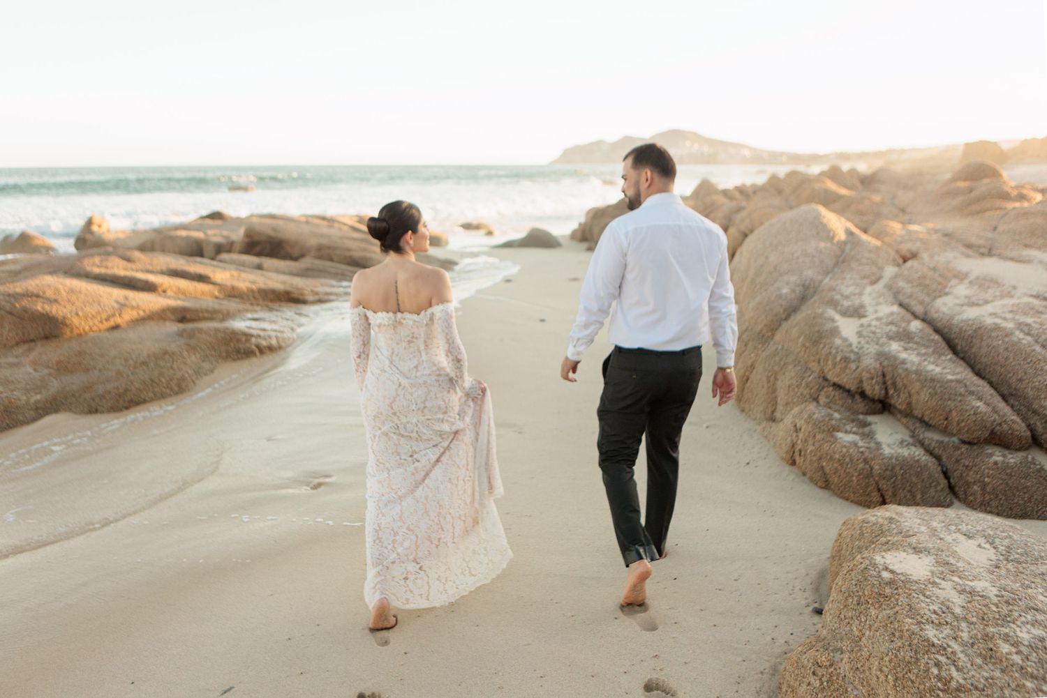 Couple in wedding attire walking hand in hand along a sandy beach with large rocks and ocean waves at sunset.