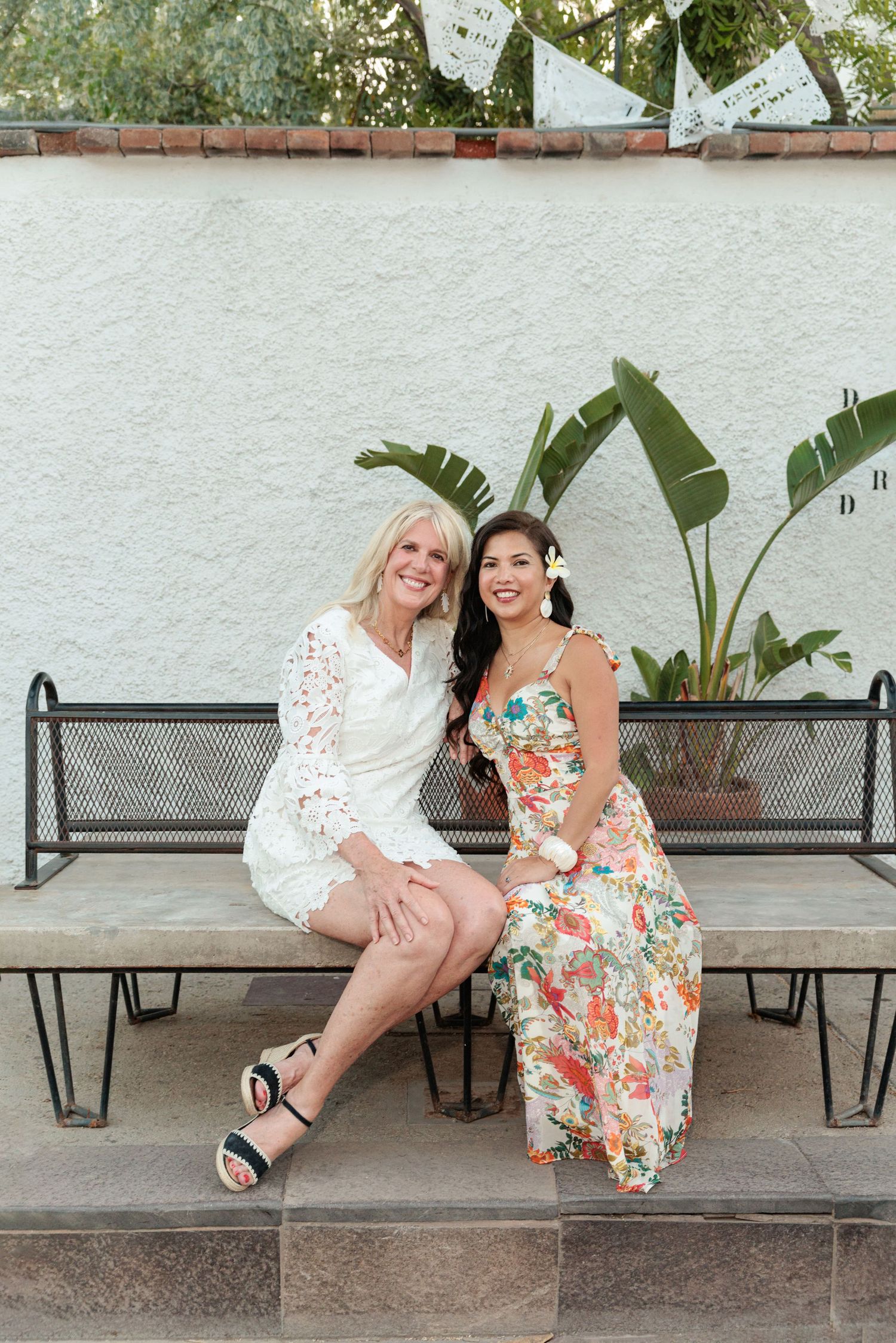 Two women smiling together on a metal bench outdoors, one in white lace dress, other in floral maxi dress.