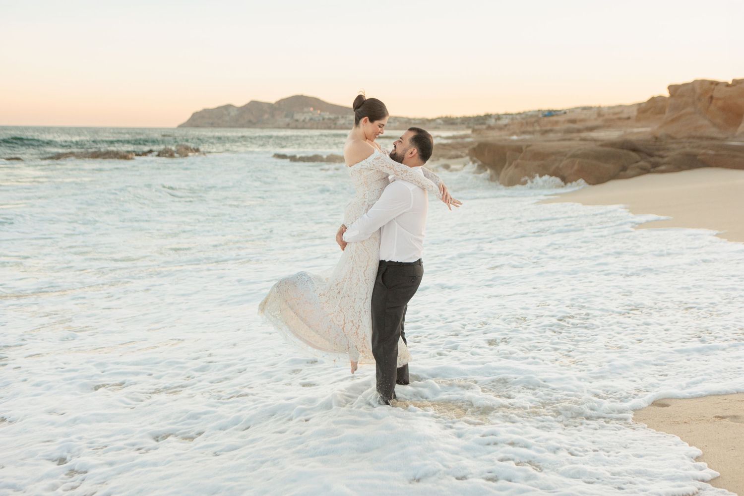 Groom joyfully lifts bride in lace gown at ocean's edge, waves crashing around them at sunset.