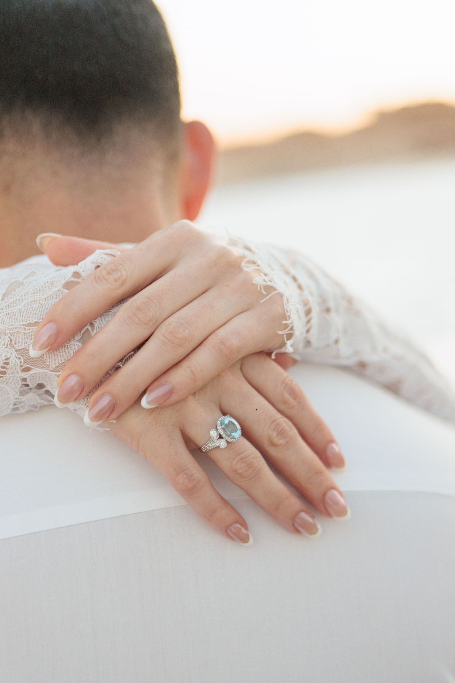 Close-up of bride's hands resting on groom's shoulder, showcasing stunning aquamarine engagement ring and lace sleeve.