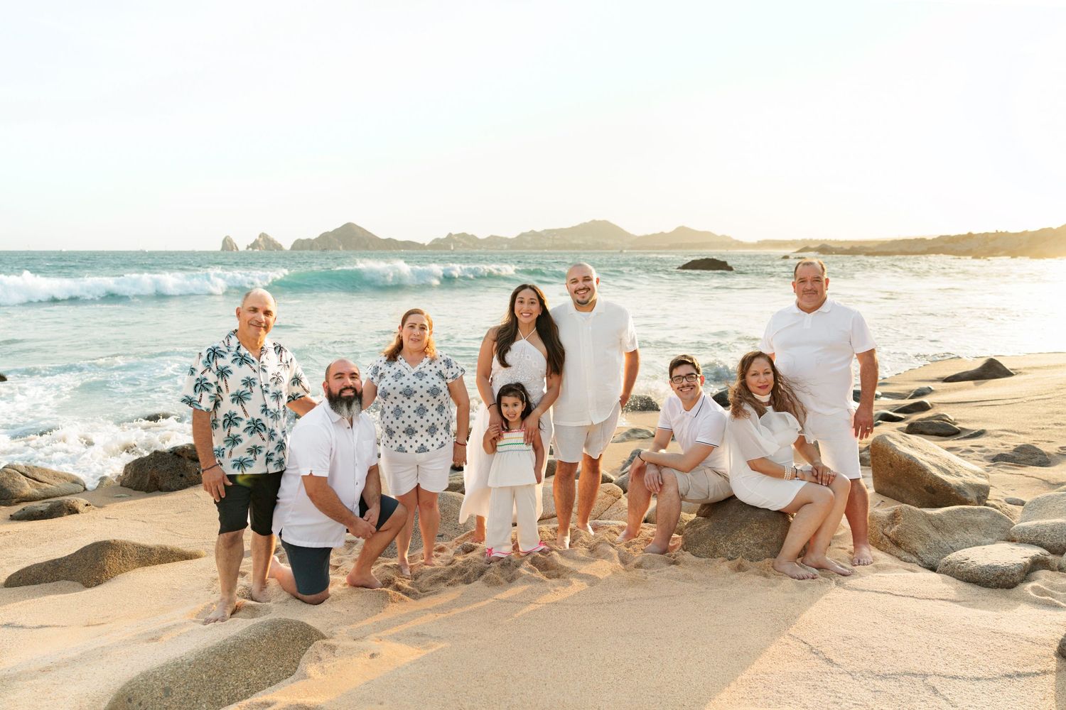 Large family group posing together on a rocky beach at sunset with ocean waves and coastal cliffs in the background.