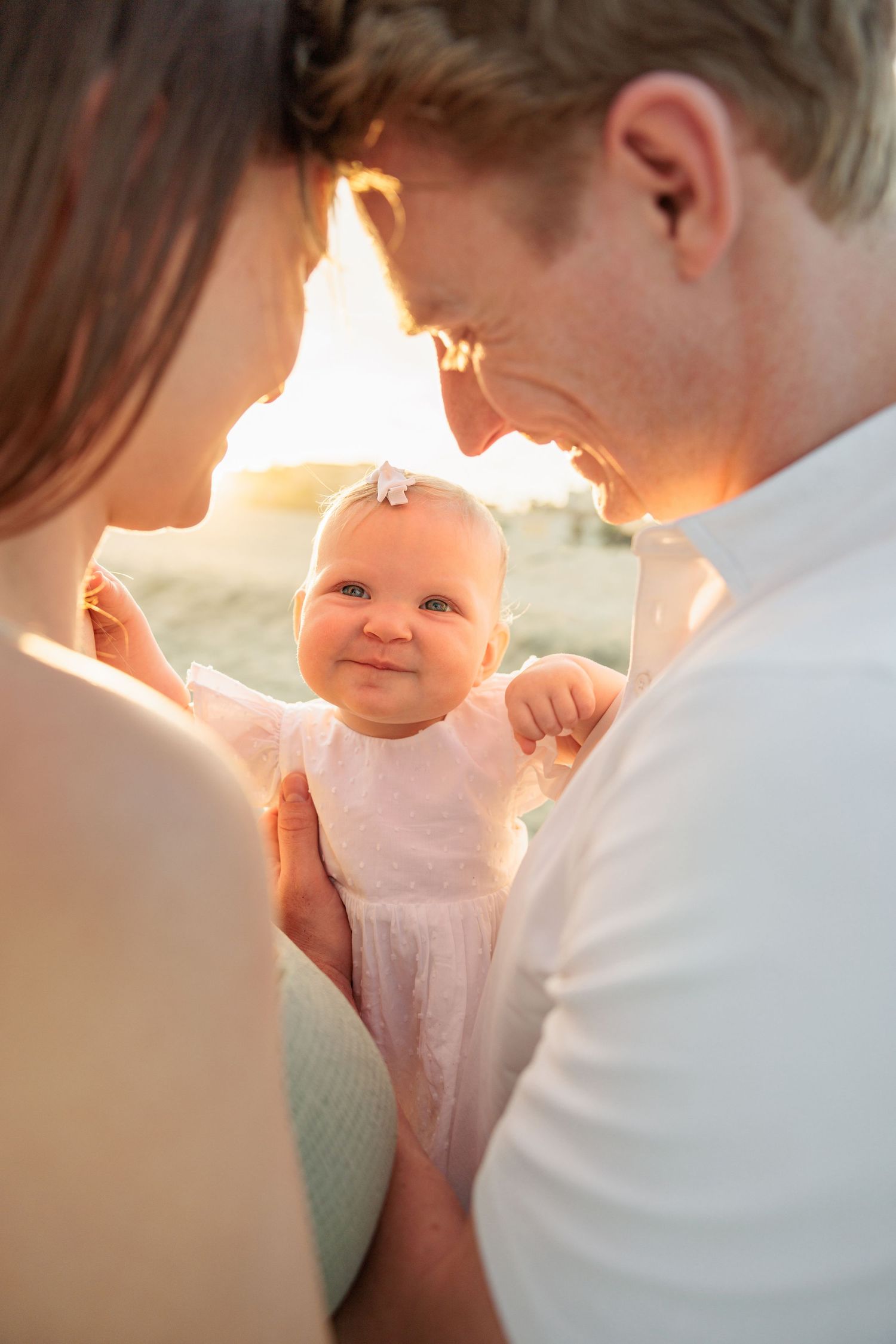 Parents holding smiling baby girl in pink dress at golden sunset, warm family portrait outdoors.