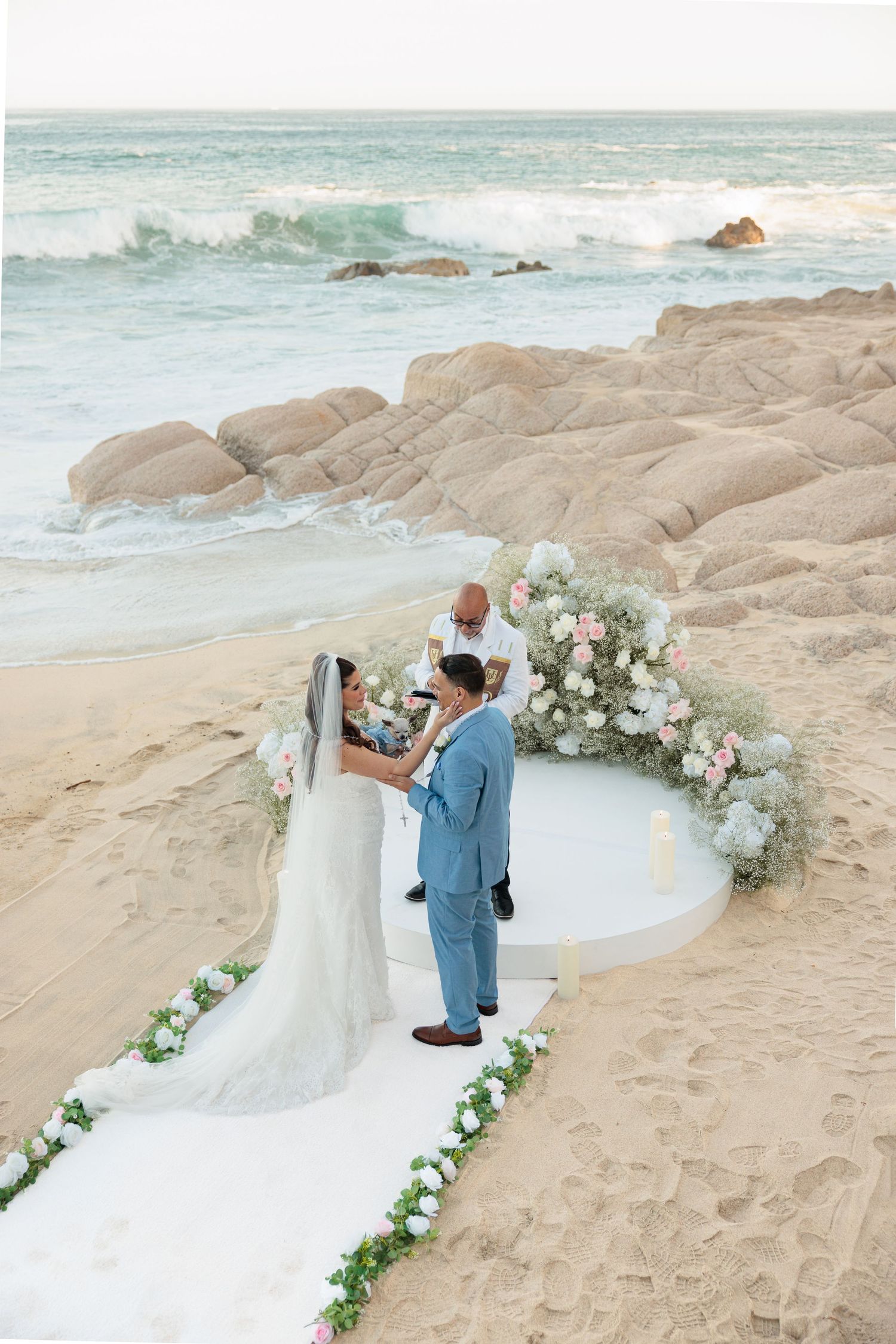 Couple exchanges vows at a floral arch on a sandy beach with rocky shores and ocean waves in the background.