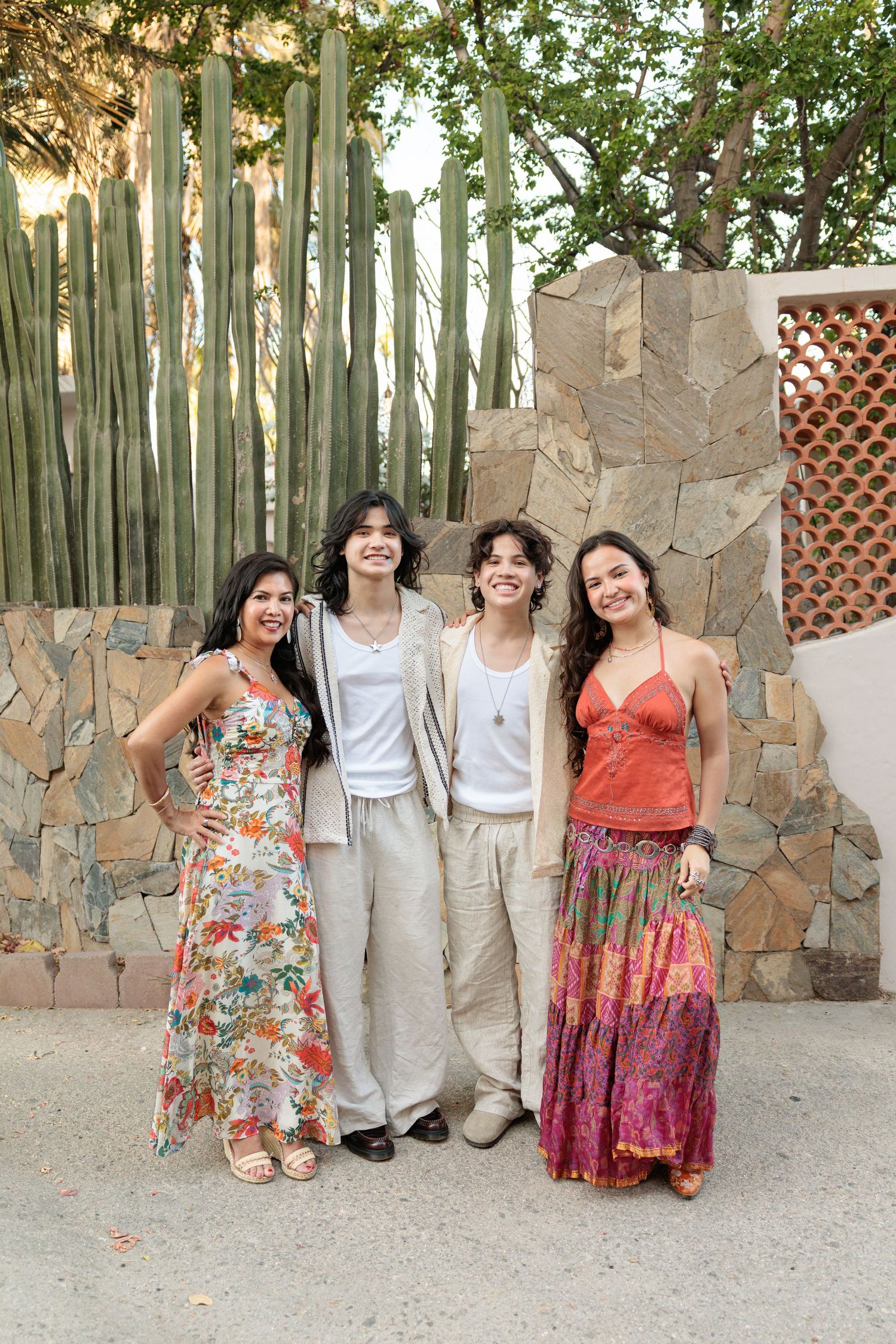Four young women smiling together outdoors near a stone wall with tall cacti, wearing colorful summer dresses and casual outfits.