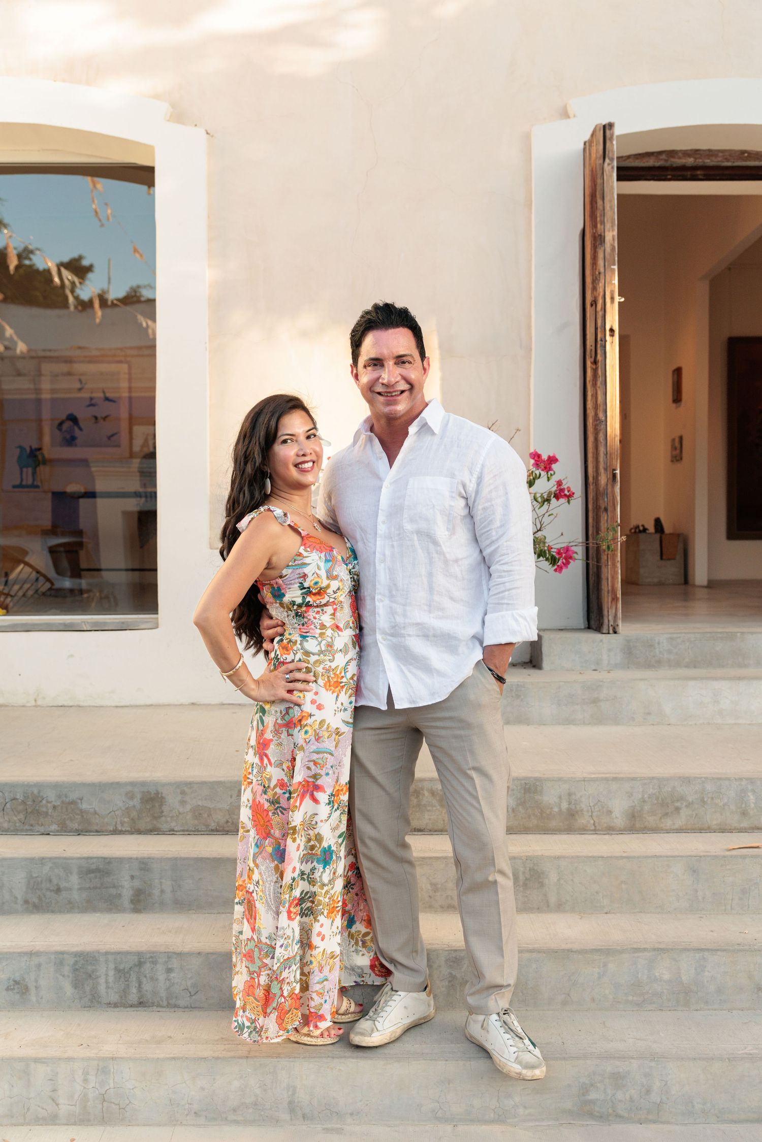 Smiling couple posing on stone steps outside a Mediterranean villa, woman in floral jumpsuit, man in white linen shirt.