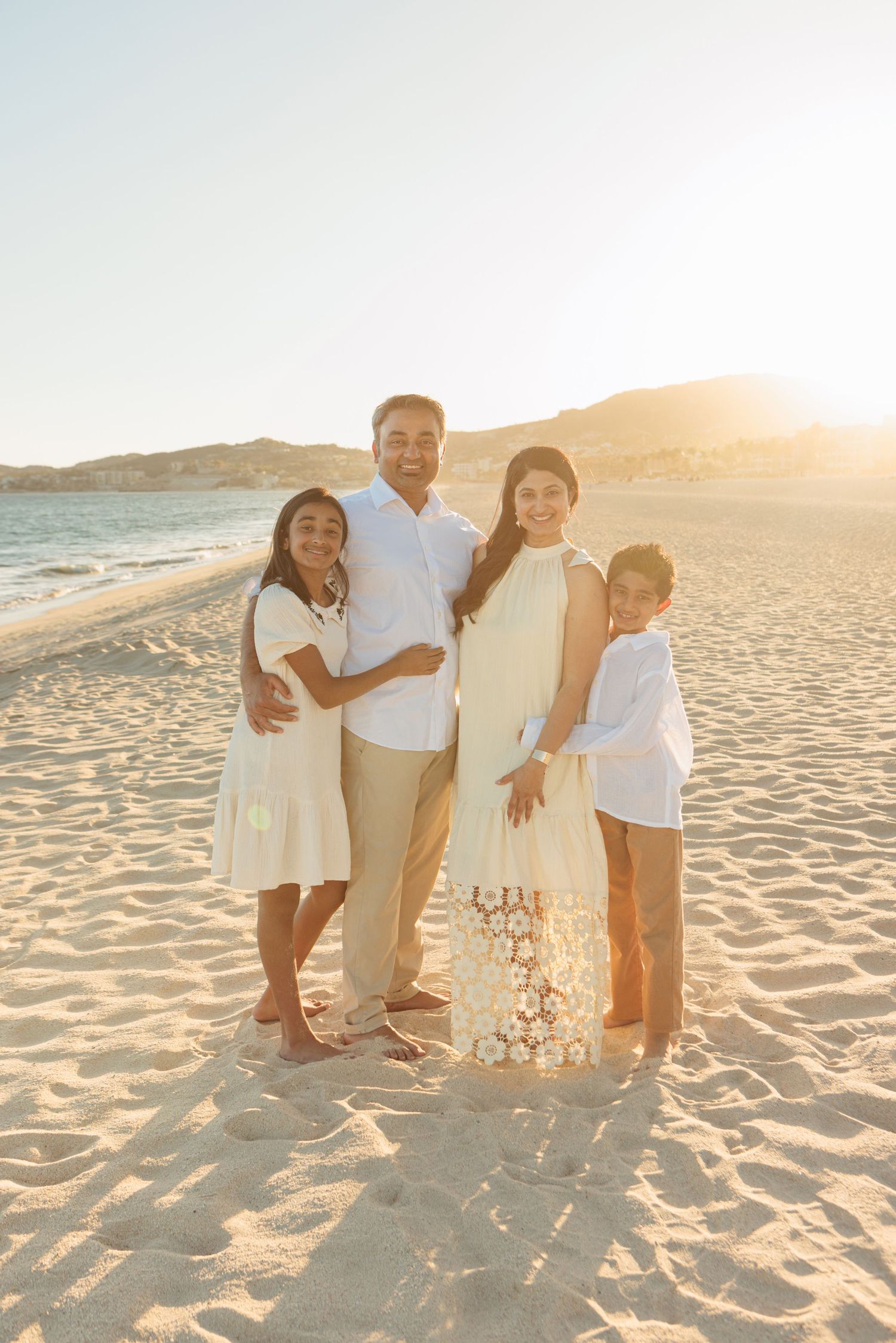 Family of four posing on sandy beach at golden sunset, wearing coordinating white and cream outfits.
