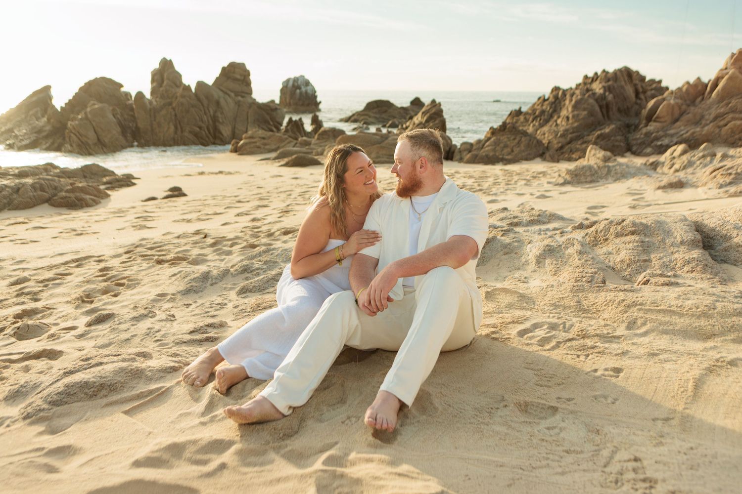 Couple sitting together on sandy beach at sunset surrounded by dramatic coastal rock formations.