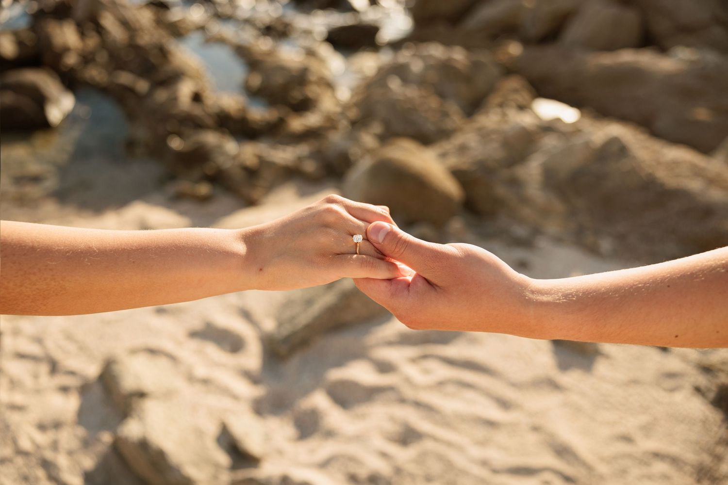 Close-up of two hands gently touching with an engagement ring on a sandy beach.