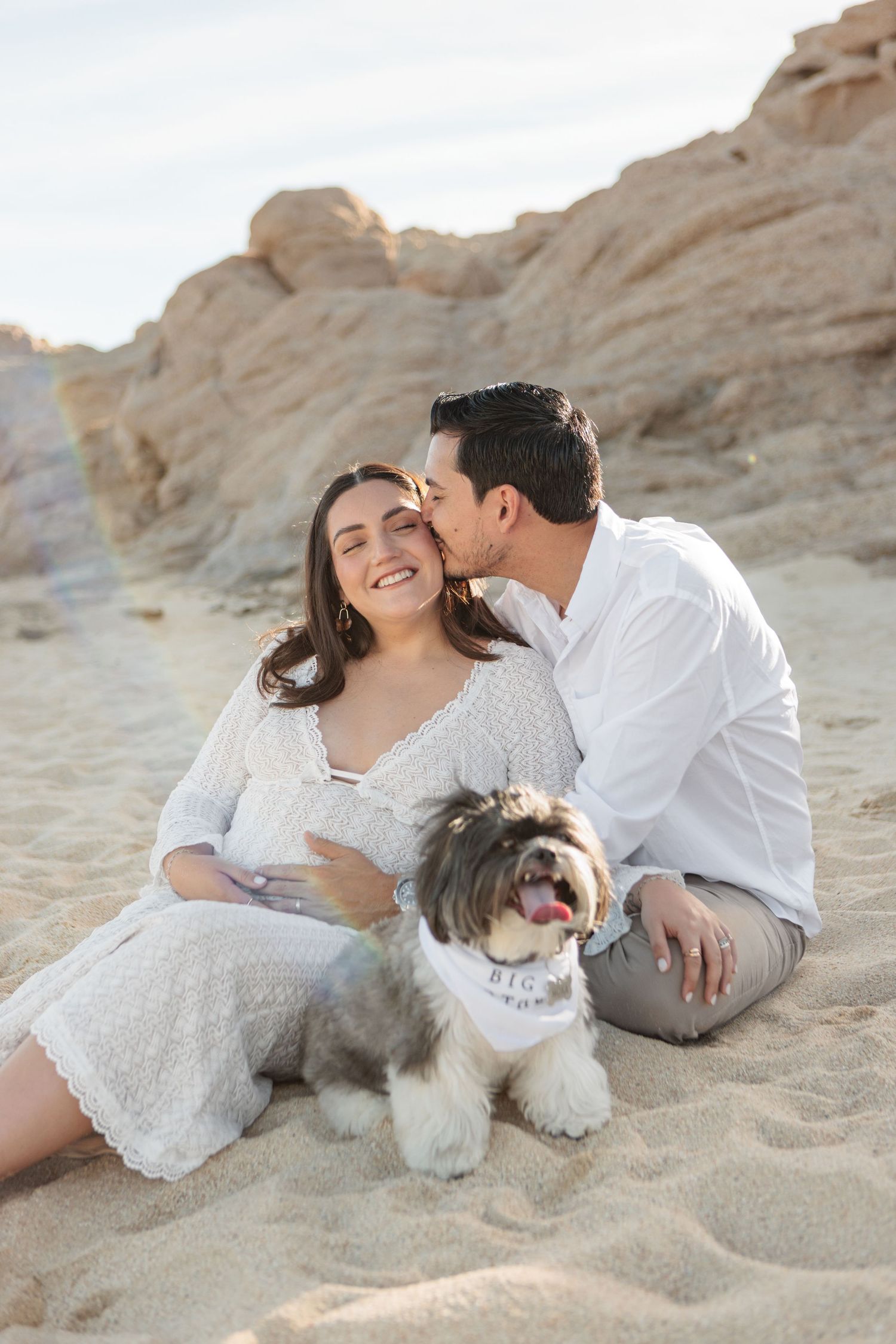 Couple sitting on sandy beach with fluffy dog wearing Big Brother bandana, man kissing woman's cheek, rocky cliffs behind them.