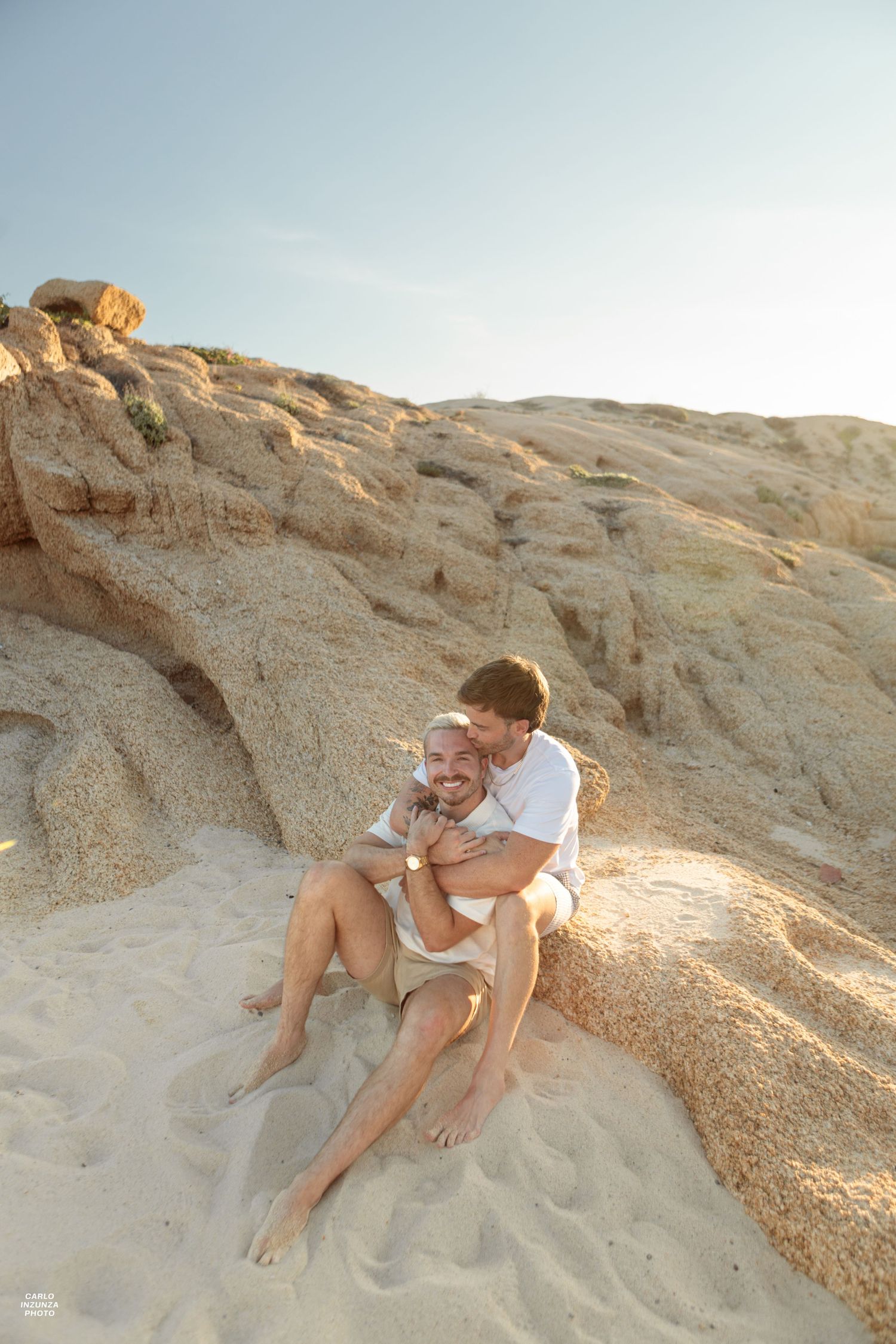 Two men embracing and laughing while sitting against sandy rock formations on beach.