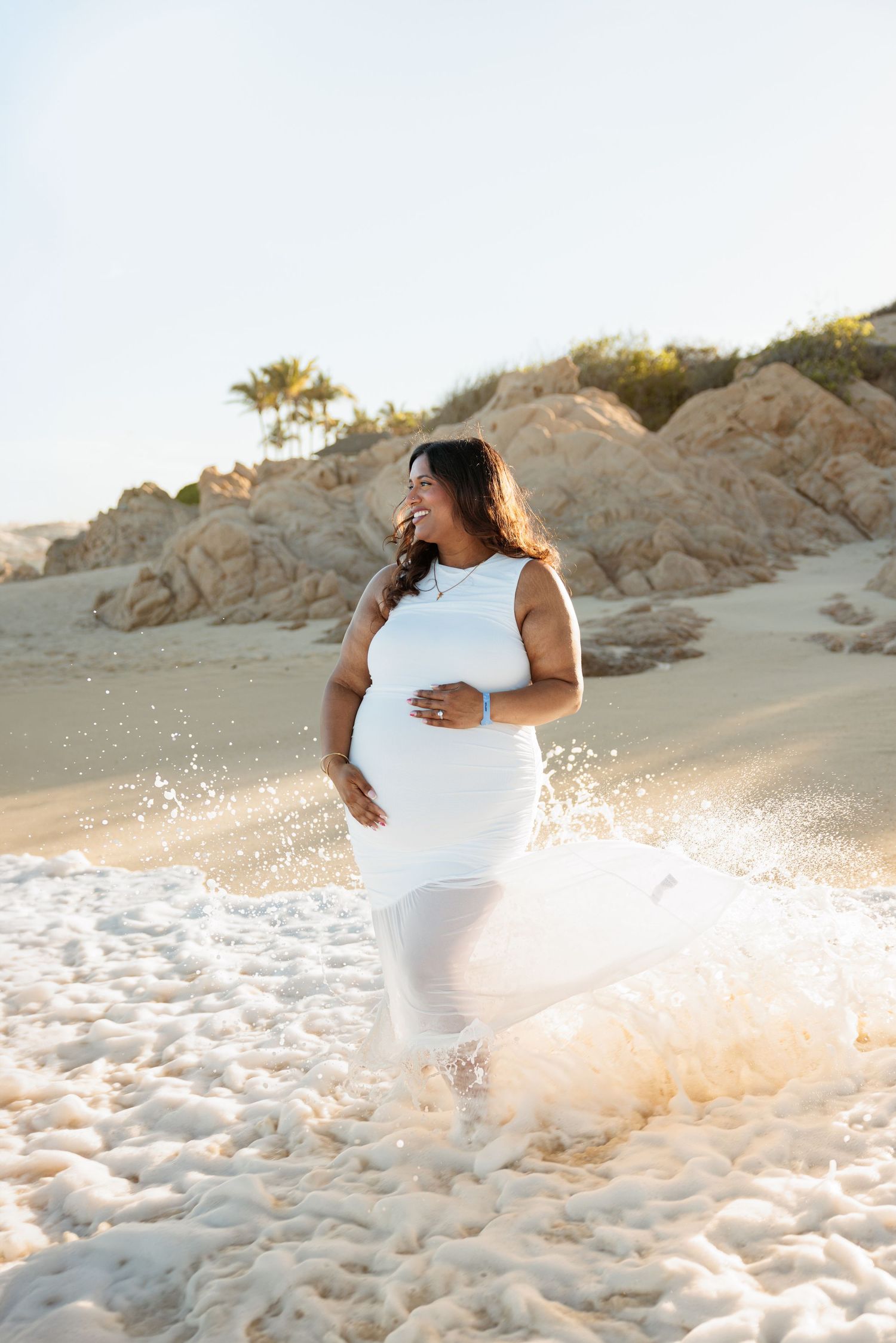 Pregnant woman in white dress standing in ocean waves on a sunny beach with rocky cliffs in background.