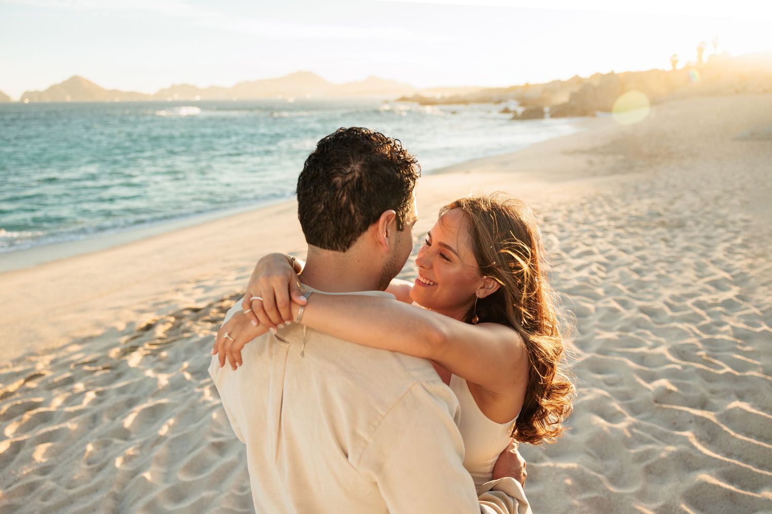 Couple embracing on a sunny beach at golden hour, smiling at each other with ocean and coastline in background.