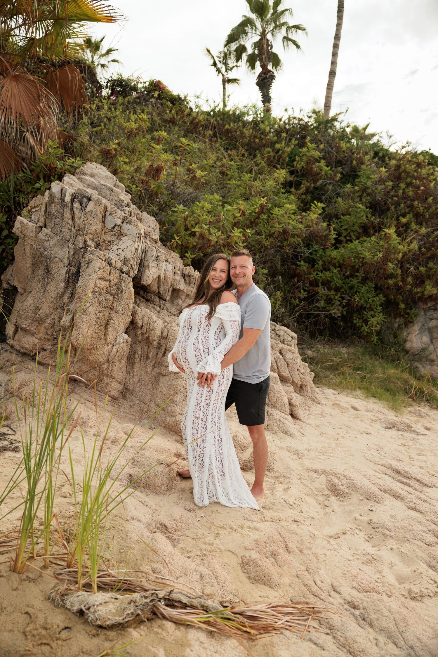 Couple posing on sandy beach with rocky cliffs and palm trees, woman in white lace dress, man in gray shirt.
