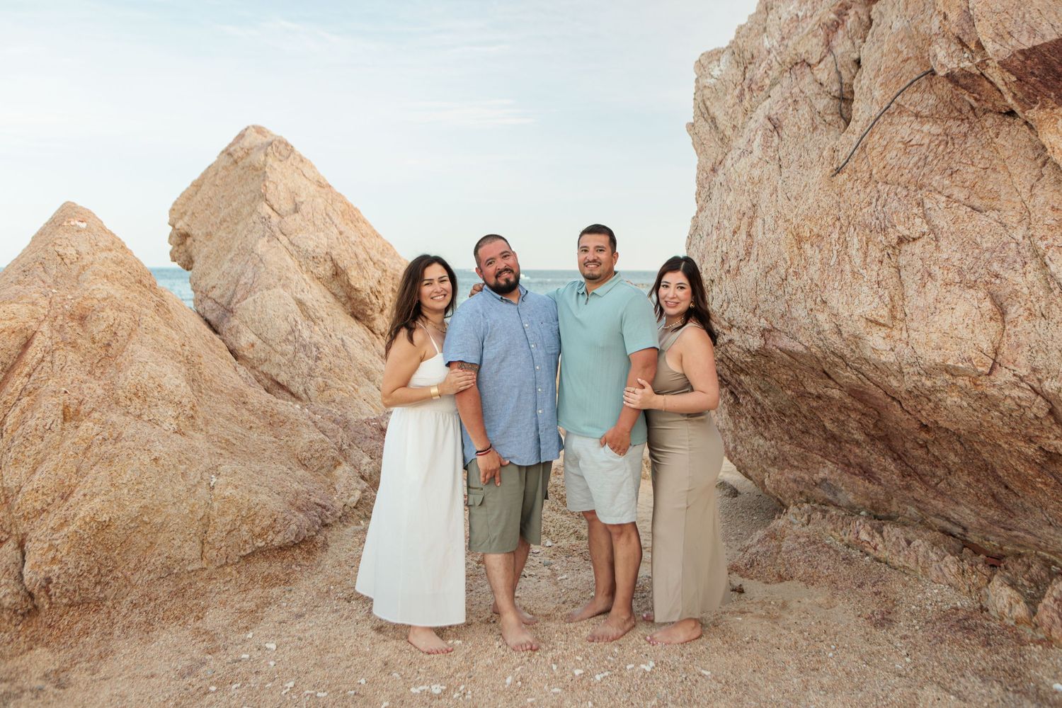 Two couples posing between large rocks on sandy beach, women in white and green dresses.