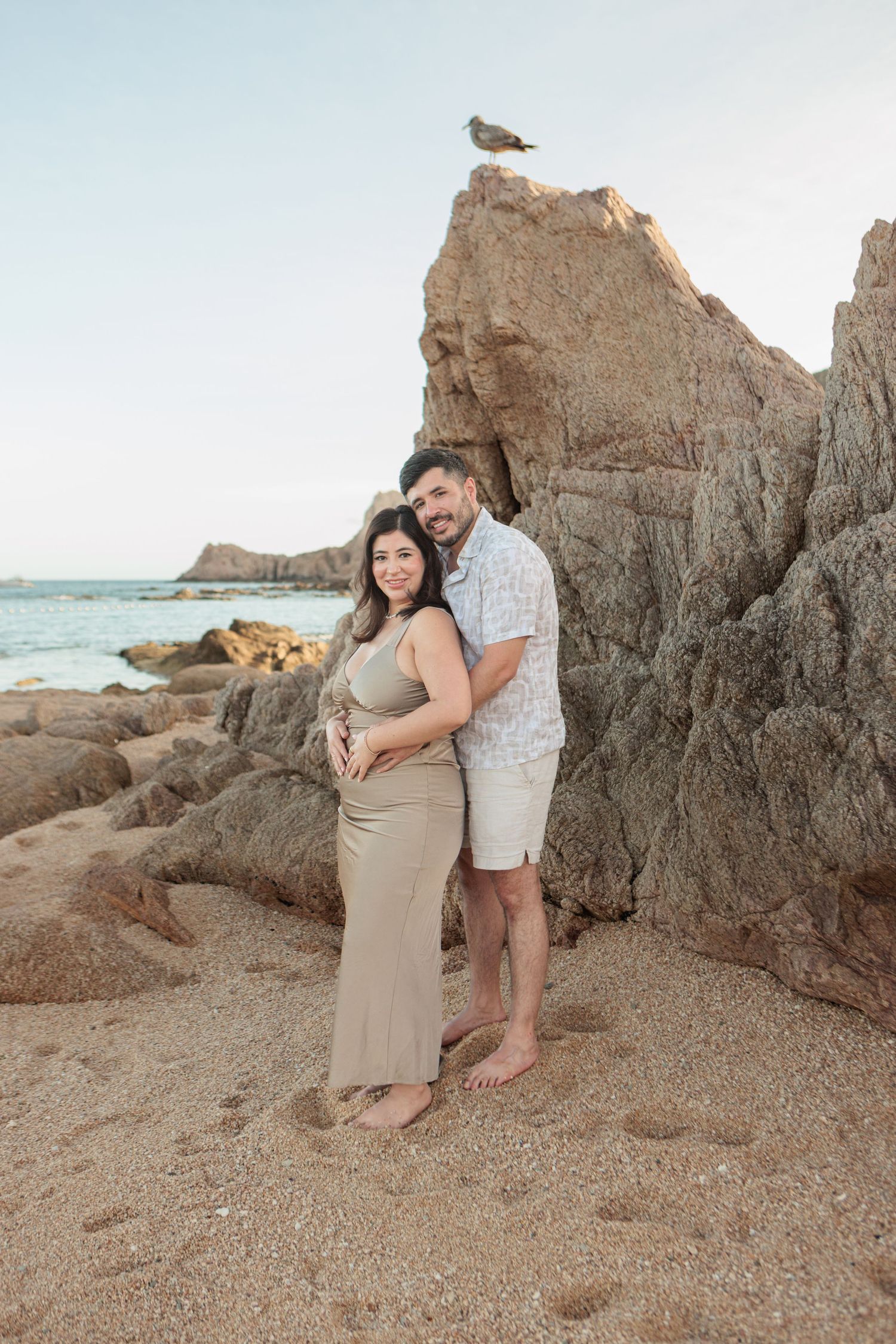 Expecting couple embracing on a rocky beach at sunset, showcasing maternity with a bird perched above.