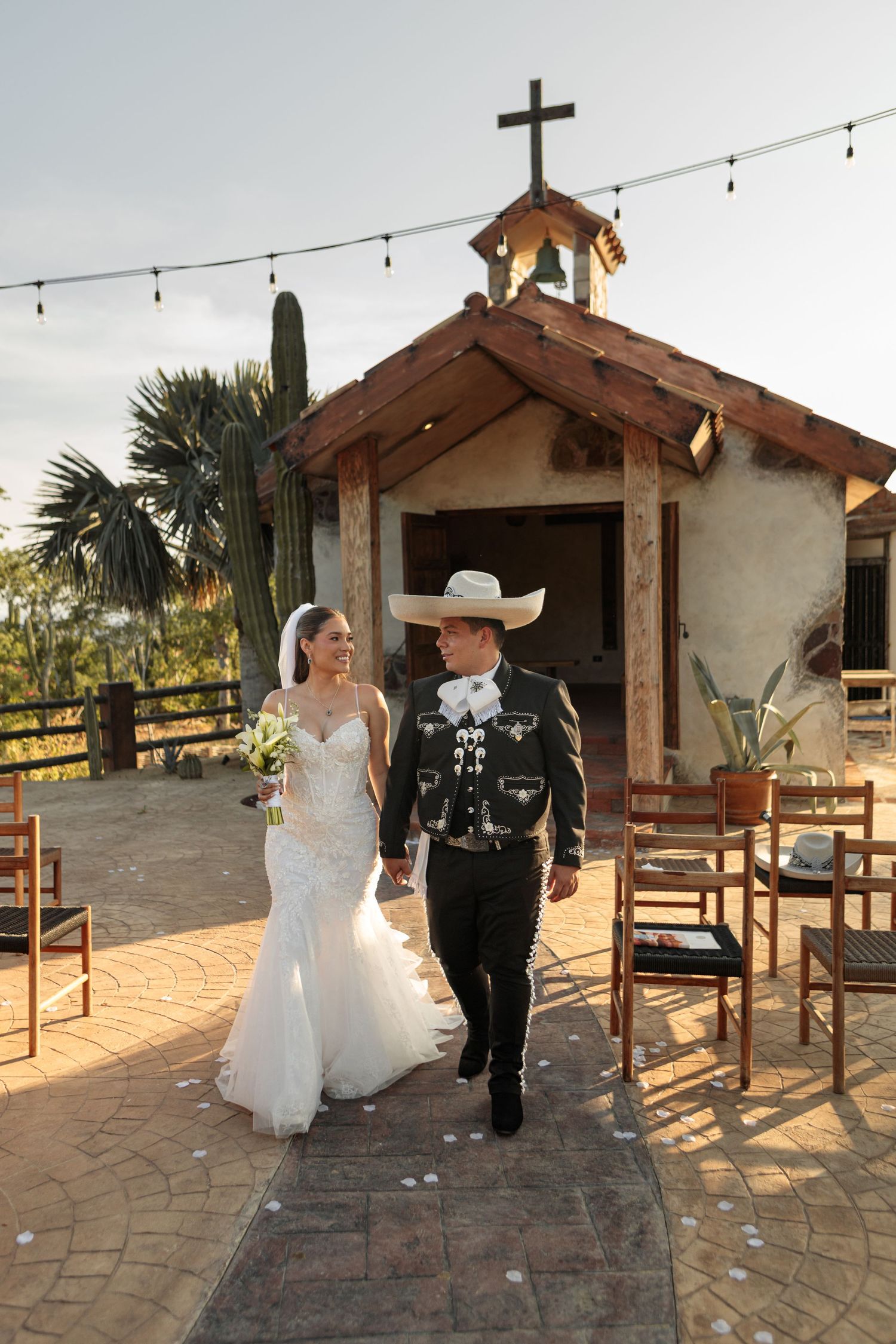 Bride and groom walking from rustic desert chapel, groom wearing traditional charro suit and cowboy hat.