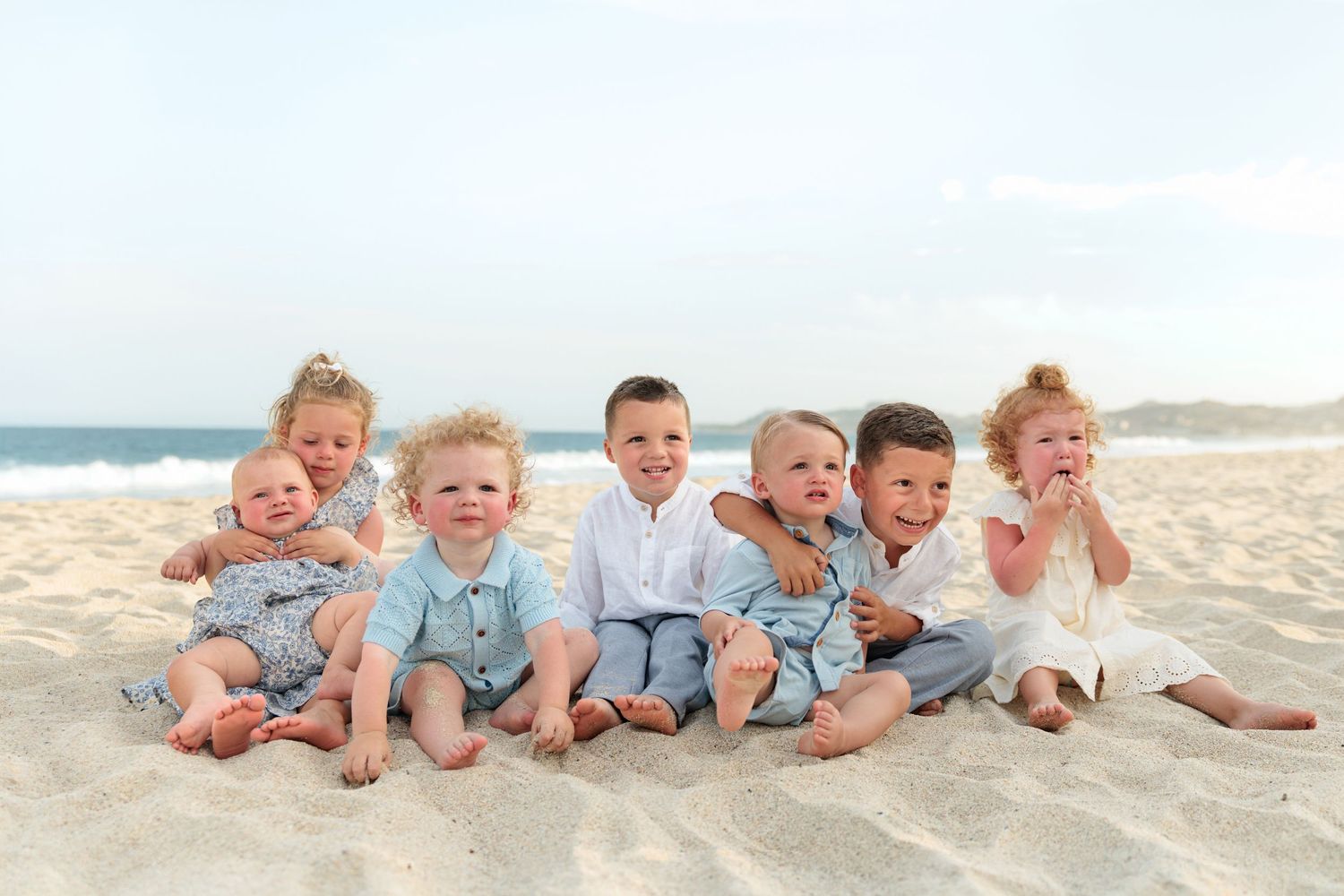 Seven young children sitting together on sandy beach, smiling for a family photo with ocean waves in background.