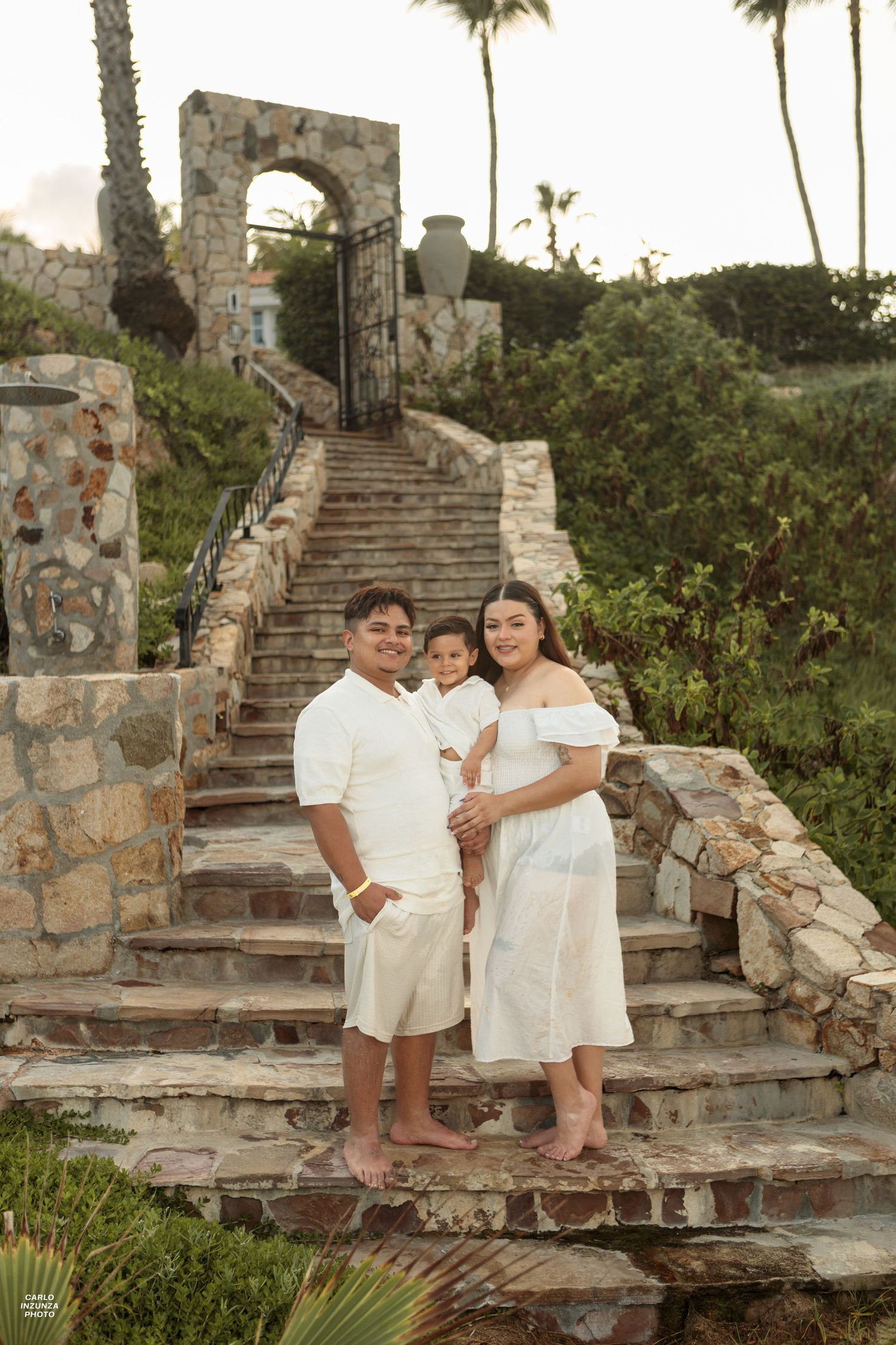 Family of three in white outfits posing on stone steps surrounded by lush tropical landscaping.