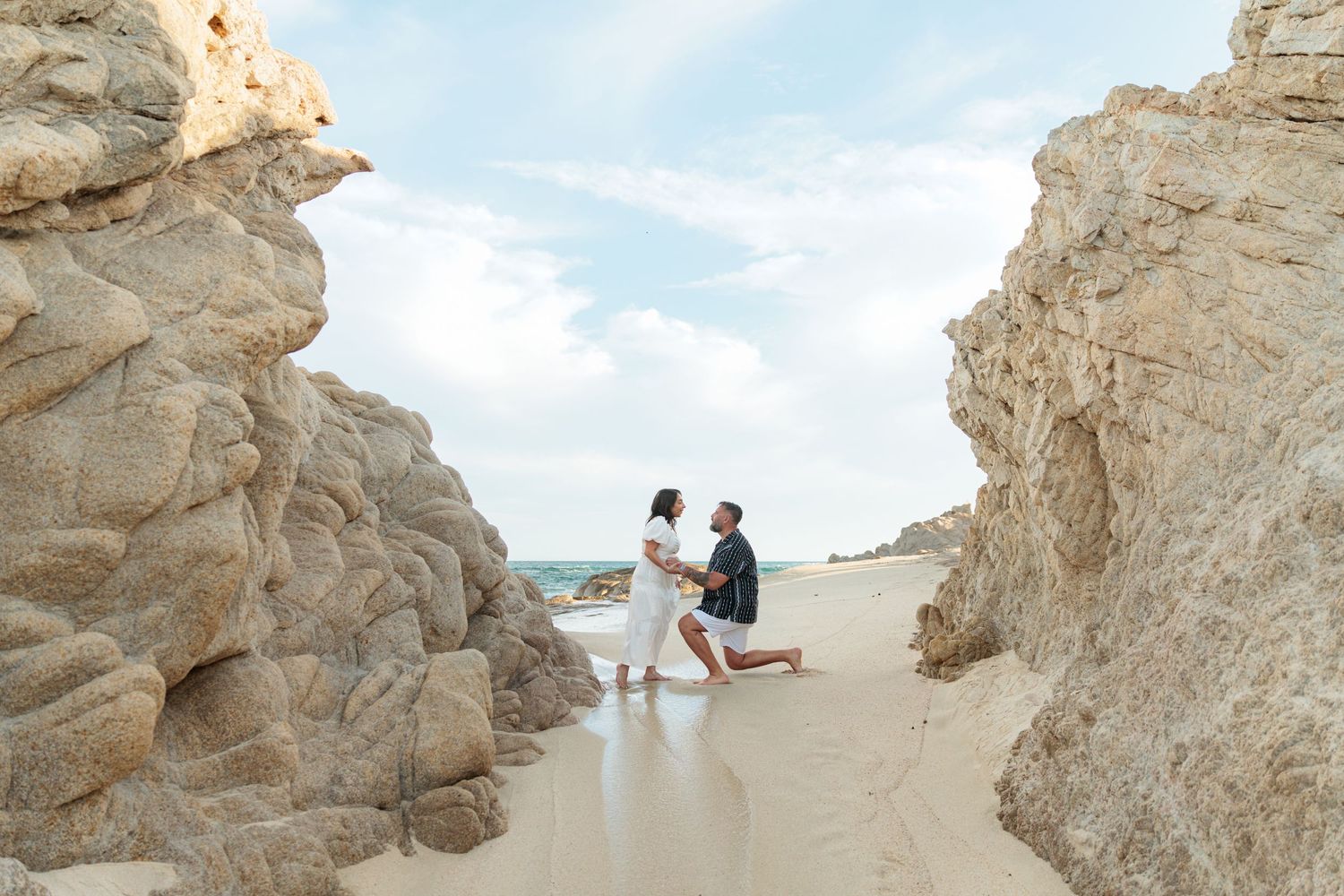 A man proposes on one knee to a woman on a sandy beach framed by large rock formations under a blue sky.