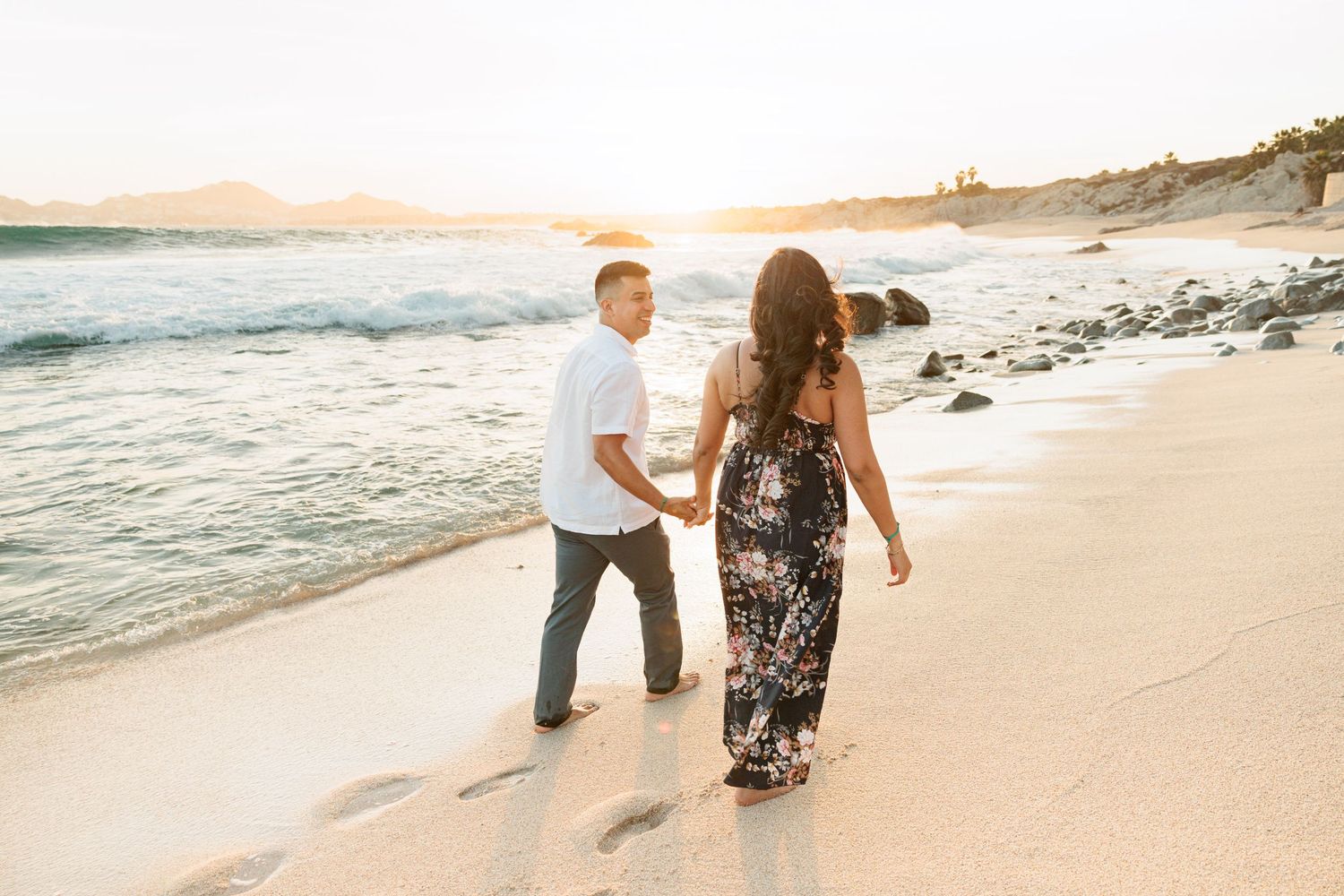 Couple holding hands walking on a sandy beach at golden sunset with ocean waves and rocky shoreline.