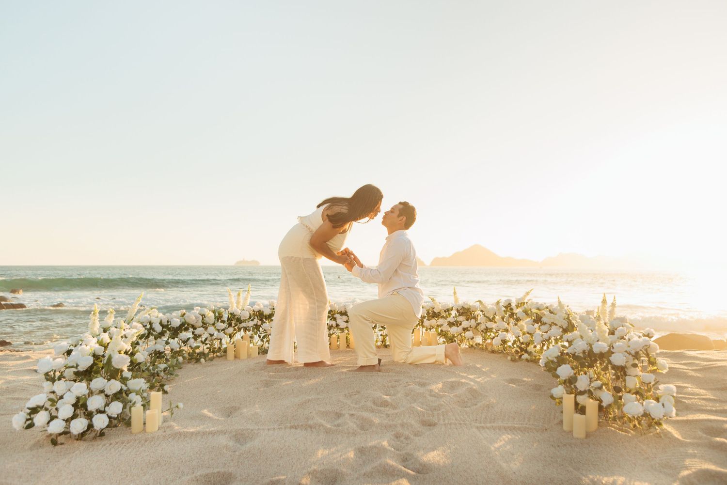 Couple kissing at sunset beach ceremony surrounded by white floral crescent arrangement with ocean in background.
