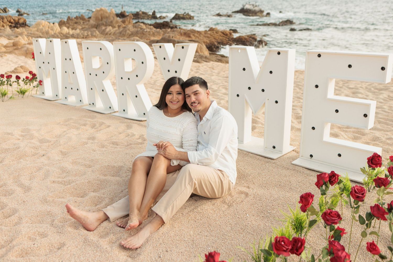 Smiling couple sitting on beach sand in front of illuminated MARRY ME marquee letters with red roses.