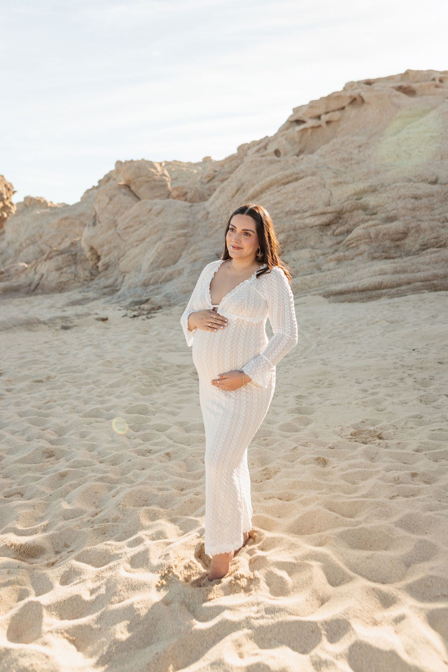 Pregnant woman in white outfit cradling baby bump on sandy beach with rocky cliffs in golden sunlight.