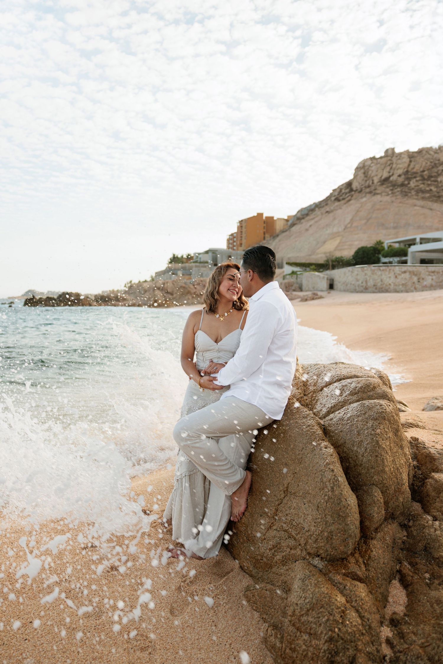 Couple in wedding attire sitting on a rock by the ocean beach with waves crashing, mountains in background.