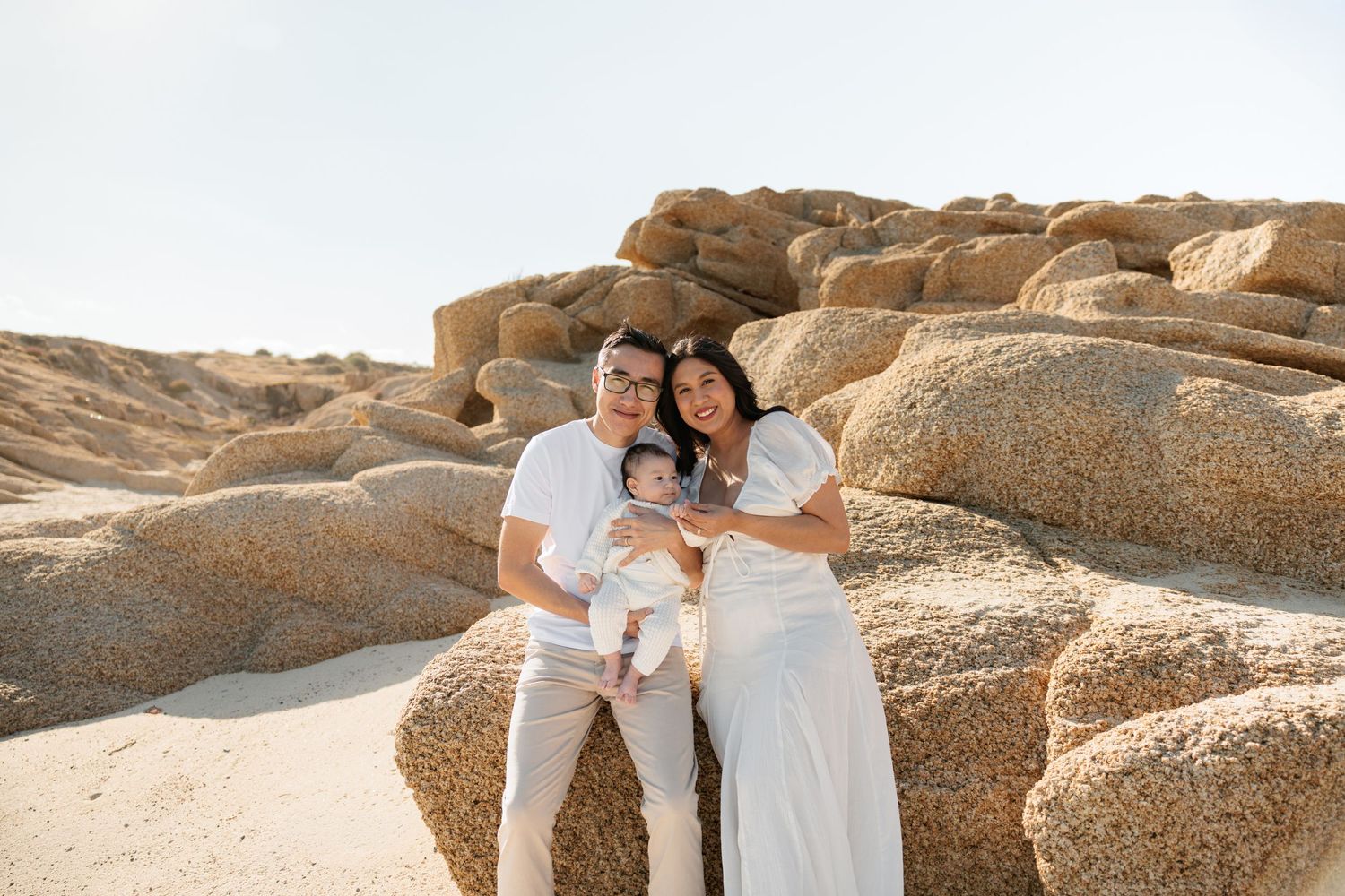 Family of three posing among sandy rock formations on a sunny beach, parents holding their baby in white outfits.