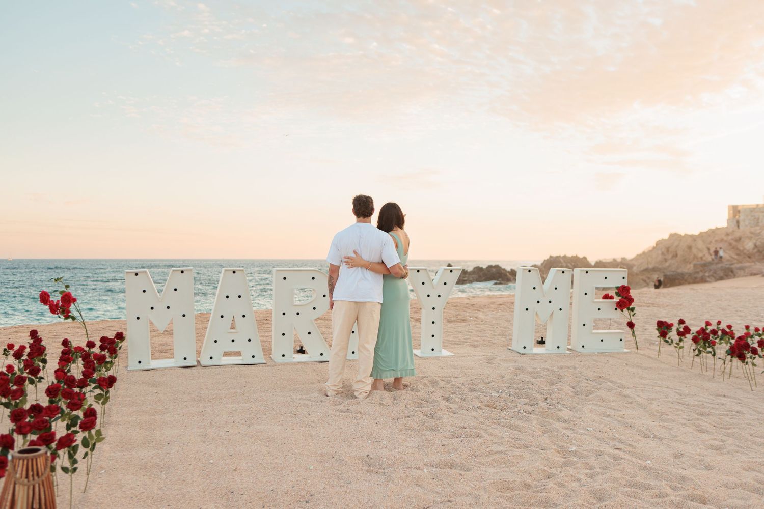 Couple standing together at beach proposal with MARRY ME marquee sign and red roses.