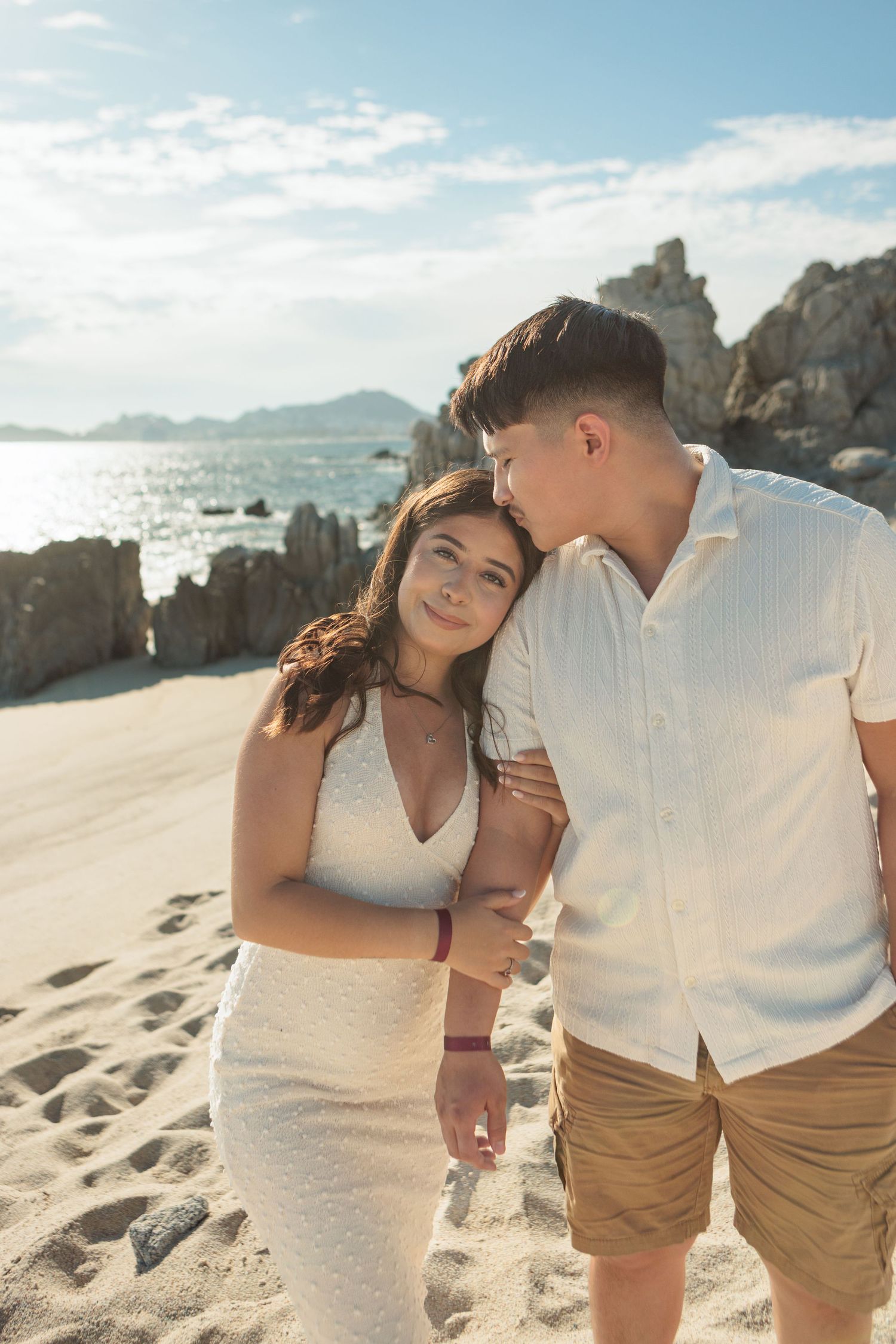 Young couple embracing on a sandy beach with rocky coastline and blue sky behind them.