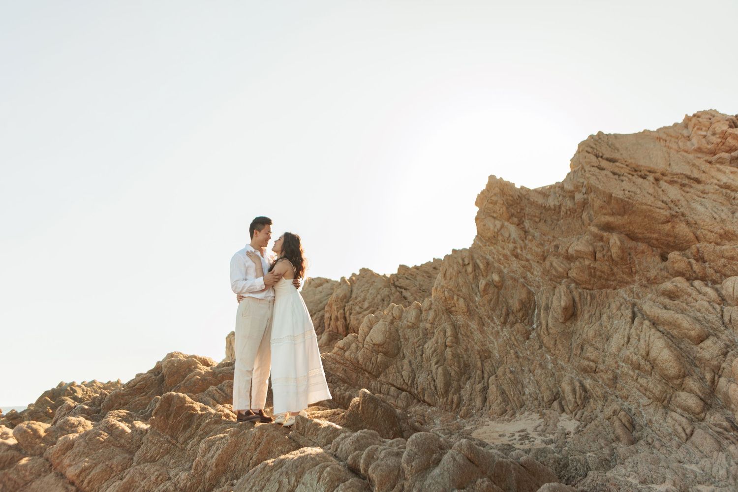 Couple embracing on top of large rocky formation with clear sky, woman in white dress, man in light outfit.