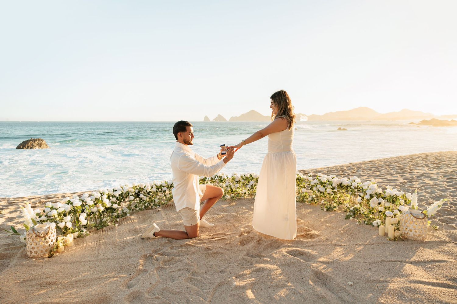 Man proposes on bended knee to woman in white dress on beach surrounded by floral arrangements at sunset.