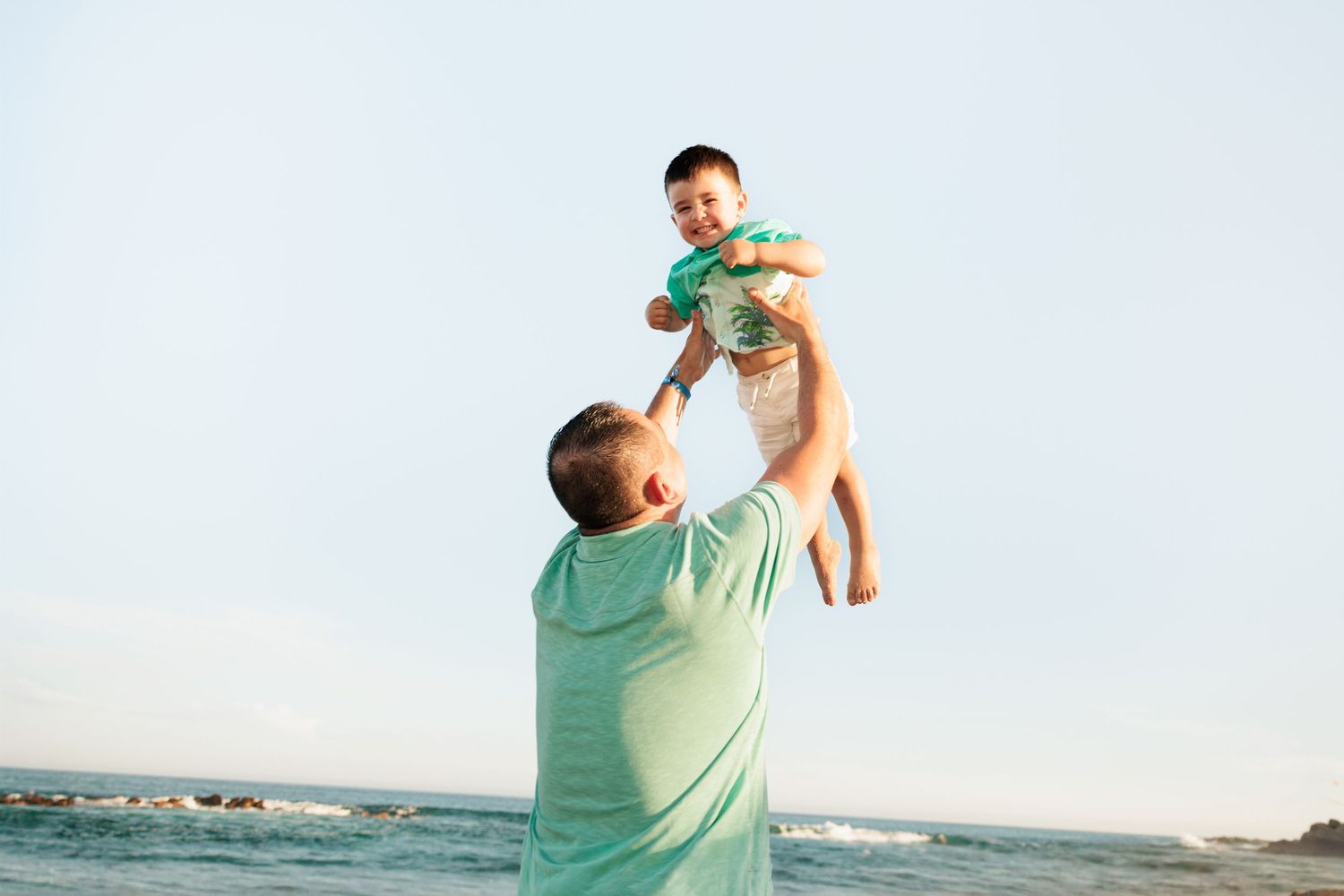 Father joyfully tossing baby in the air at the beach on a sunny day.