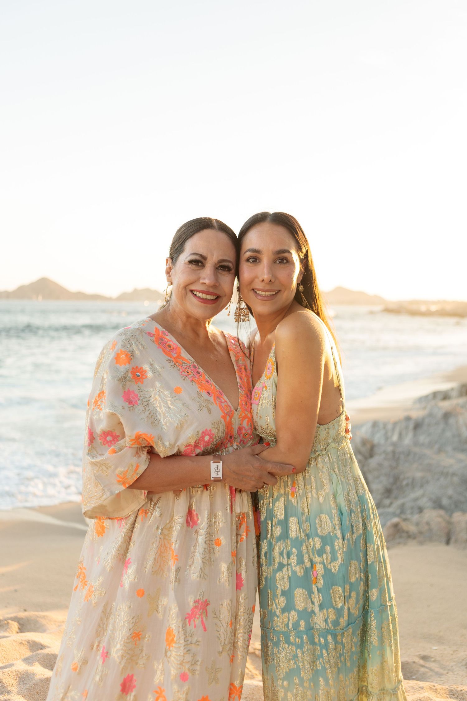 Two smiling women in elegant floral and pastel maxi dresses embracing on a beach at sunset.