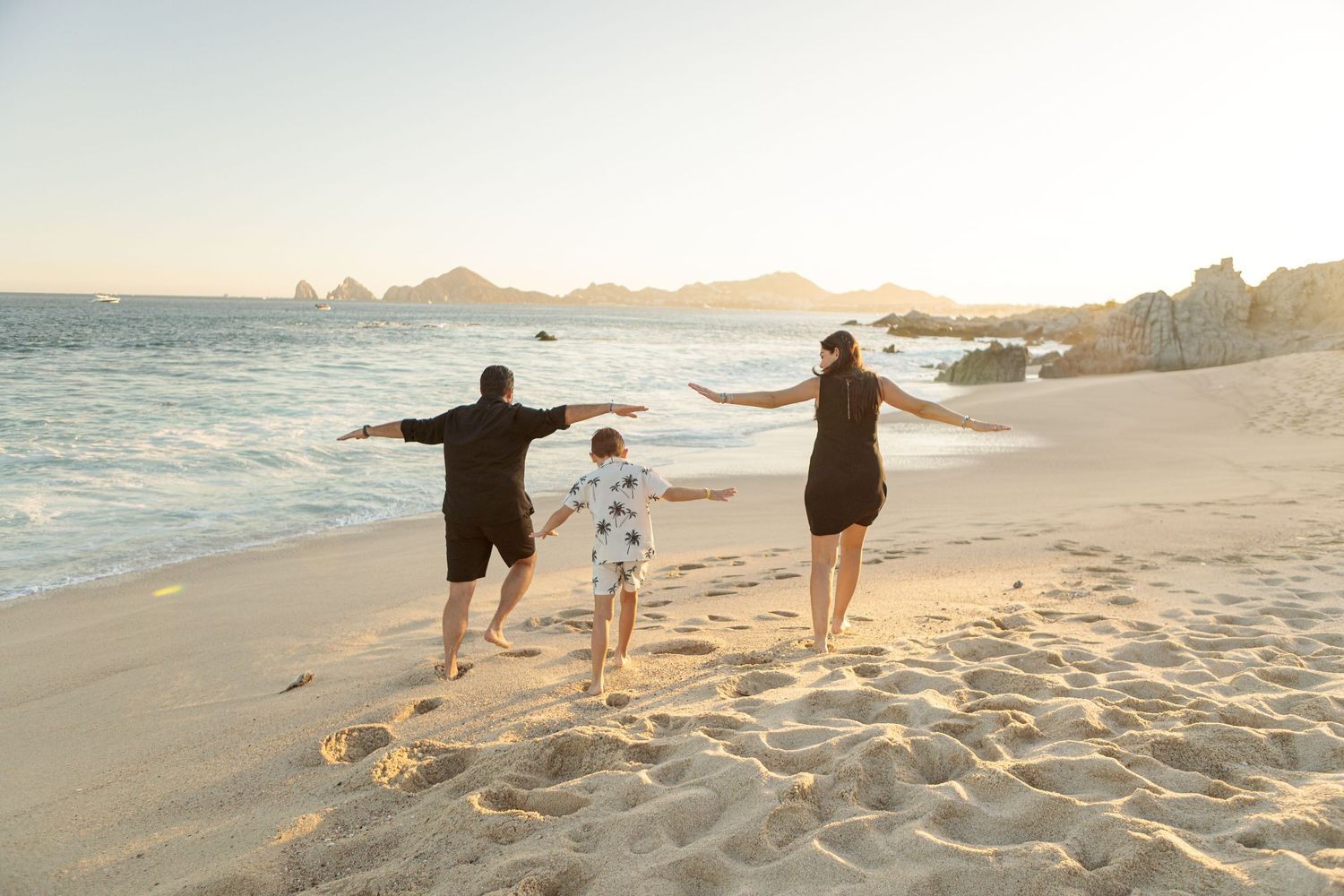 Family of three walking hand-in-hand on a sandy beach at sunset with rocky coastline in background.