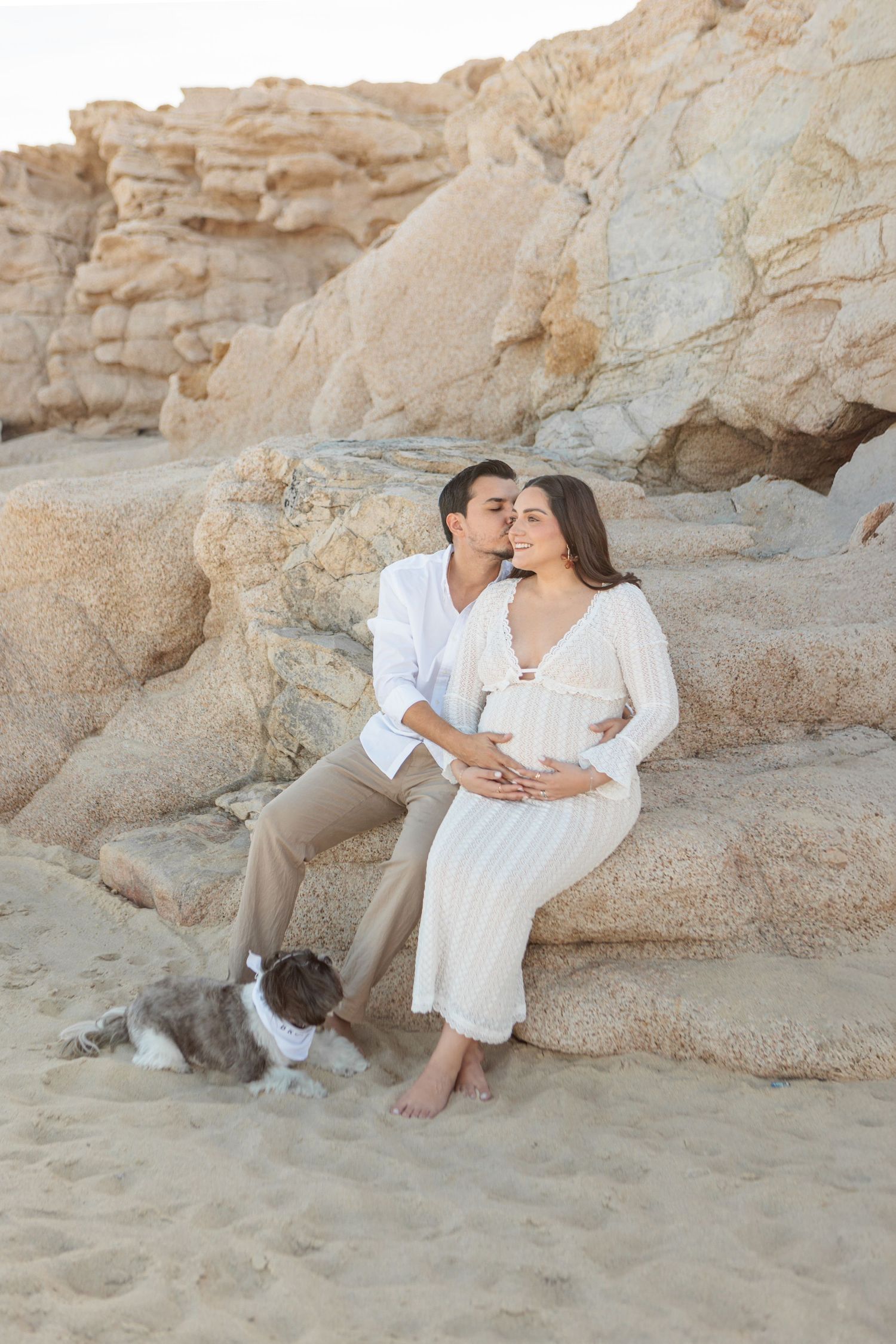 Couple sitting on beach rocks, man kissing pregnant woman in white dress, with a dog resting on sand nearby.
